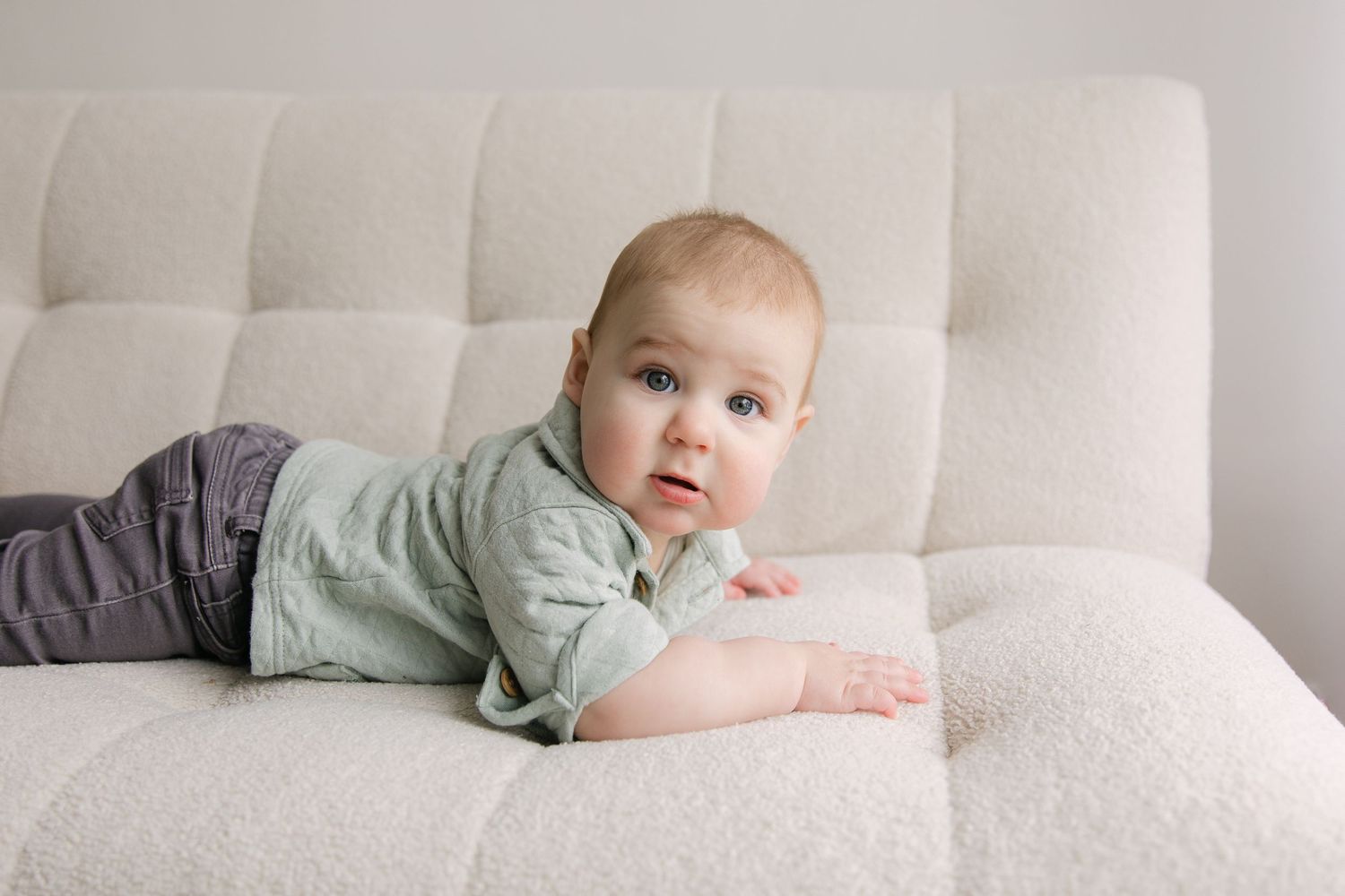 A cute baby in a green outfit lying on their tummy on a beige sofa, looking up with bright blue eyes.