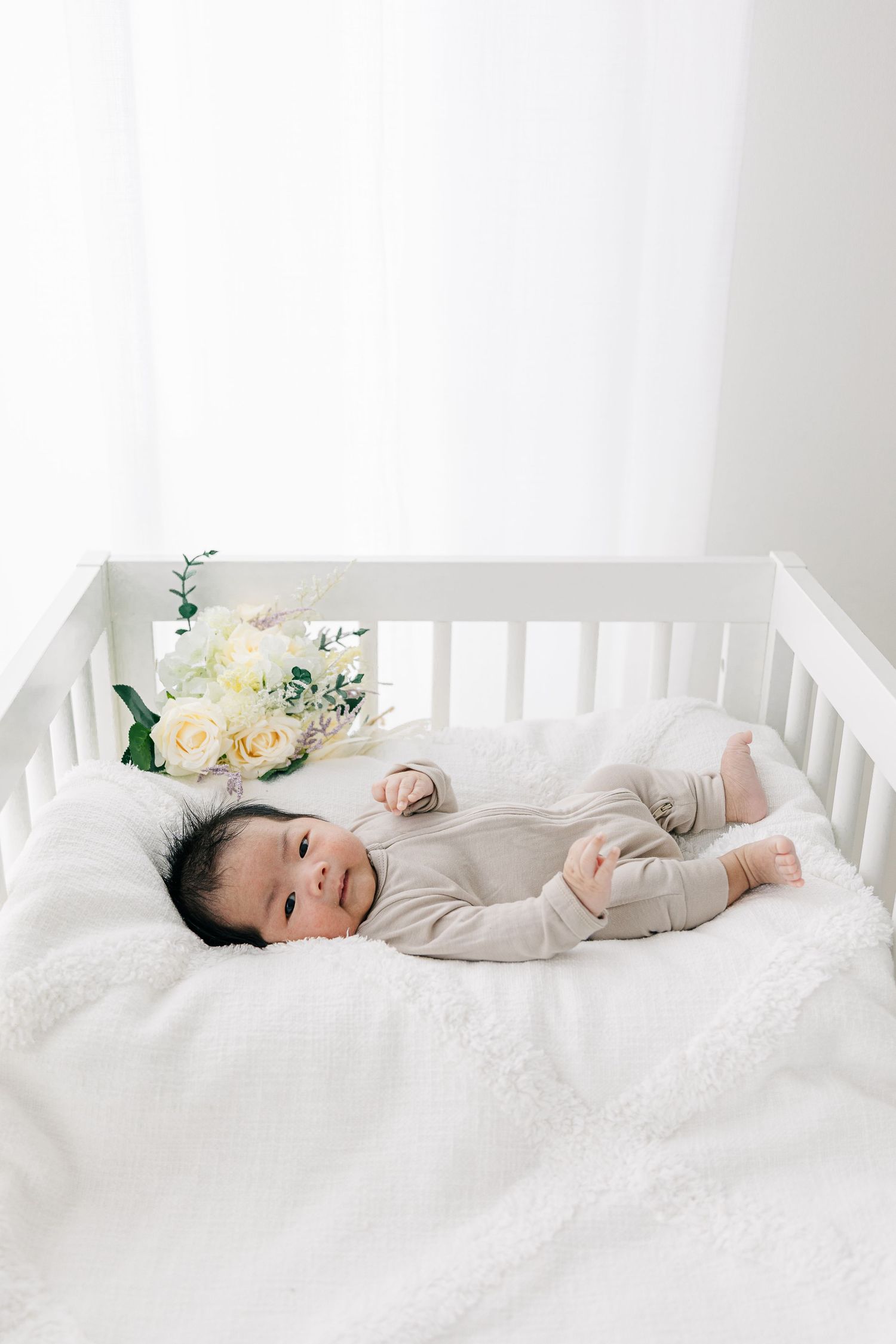 Baby in light gray onesie lying in white crib with floral decorations.