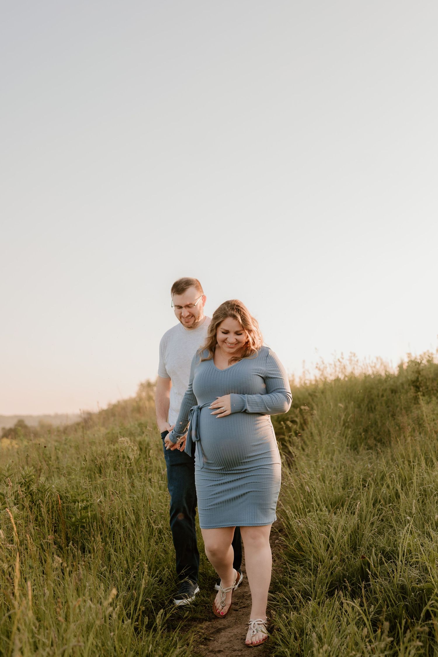 Couple walking together through tall grass during sunset maternity photoshoot.
