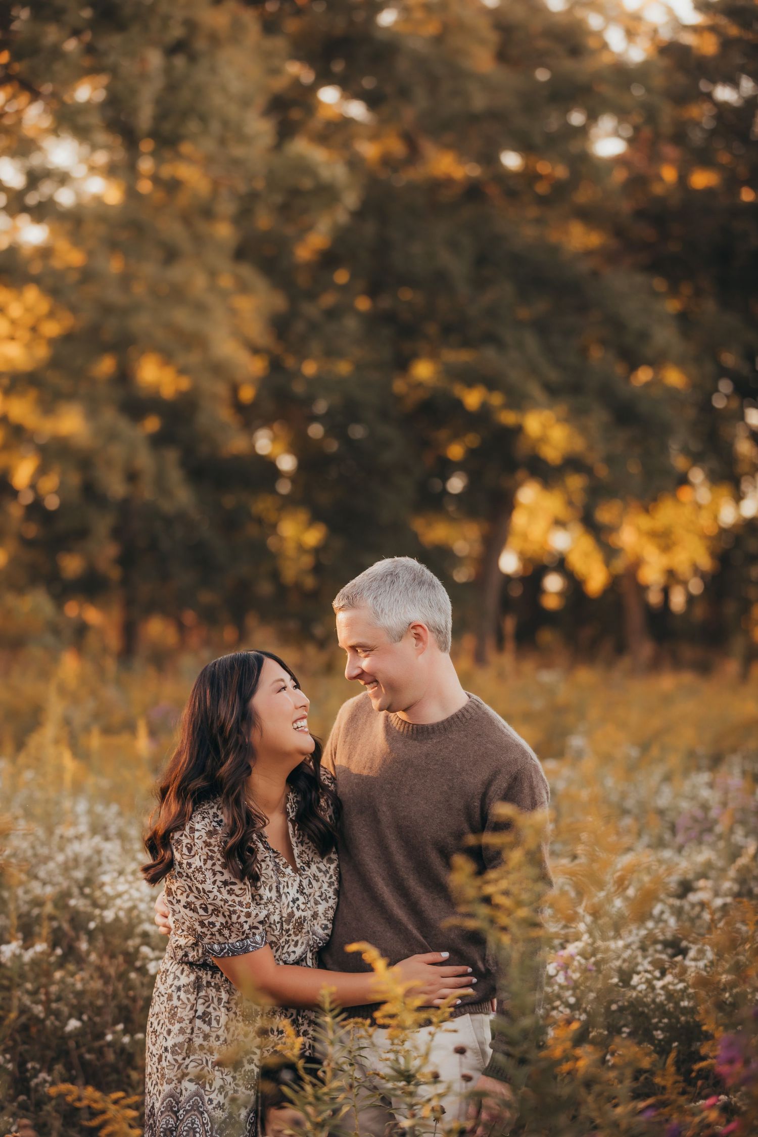 Couple laughing together in a golden autumn field surrounded by colorful fall foliage during a romantic outdoor photo session.