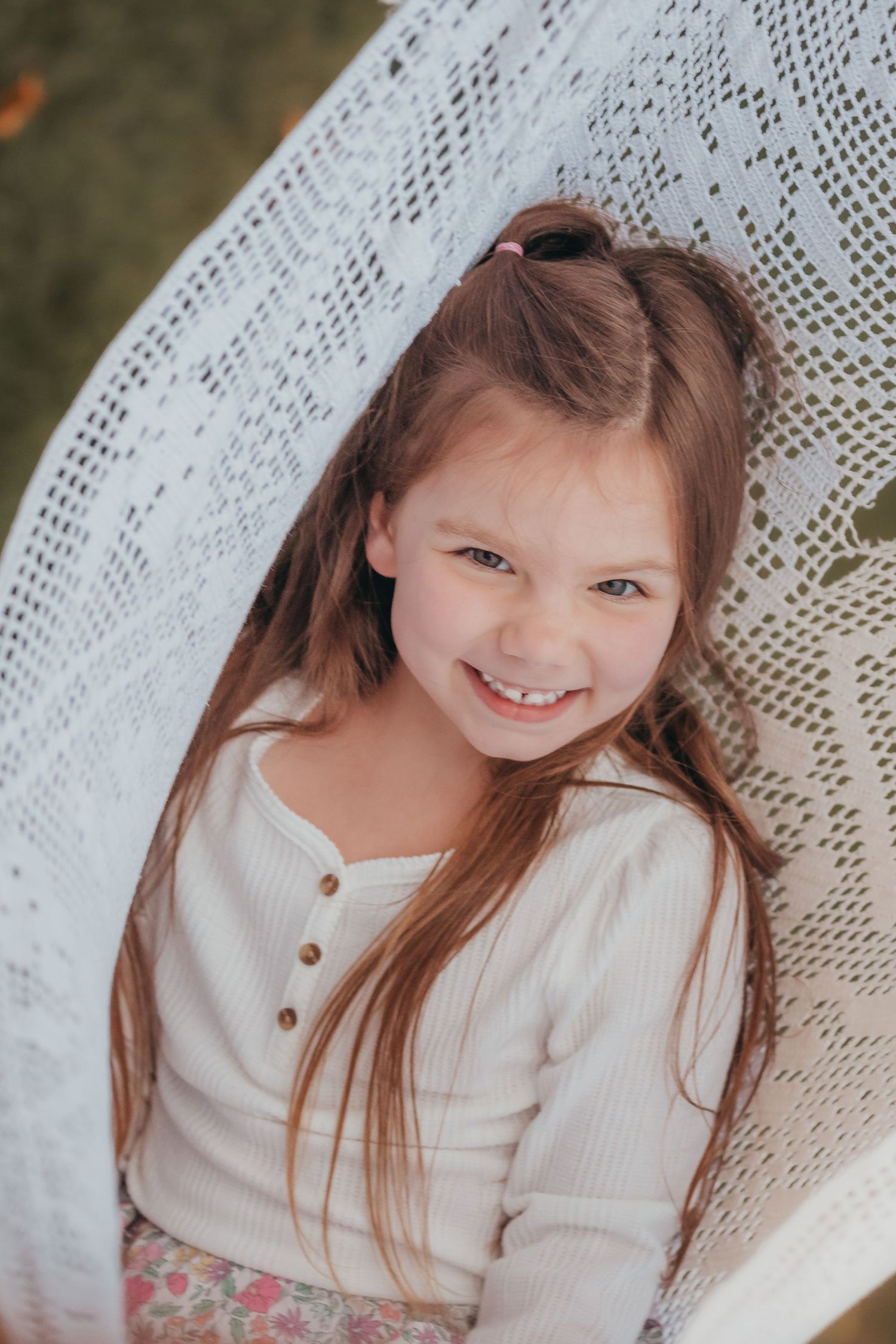 A portrait of a young child with long brown hair wearing a white sweater against a lace backdrop, smiling warmly.