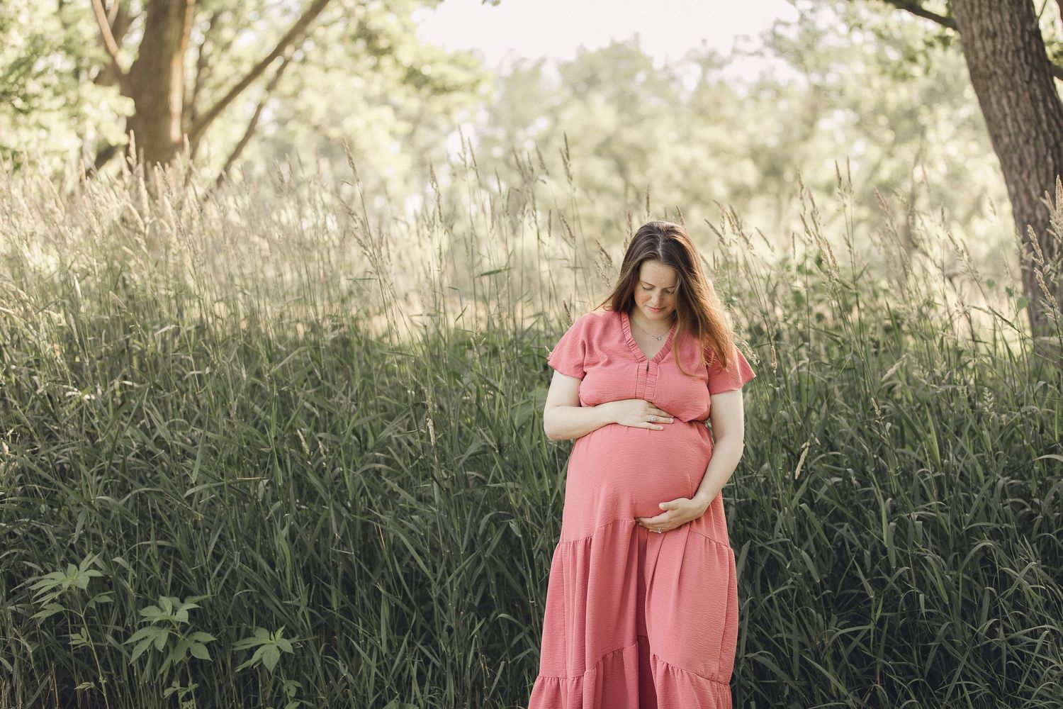 A pregnant woman in coral dress stands alone in tall grass during golden hour.