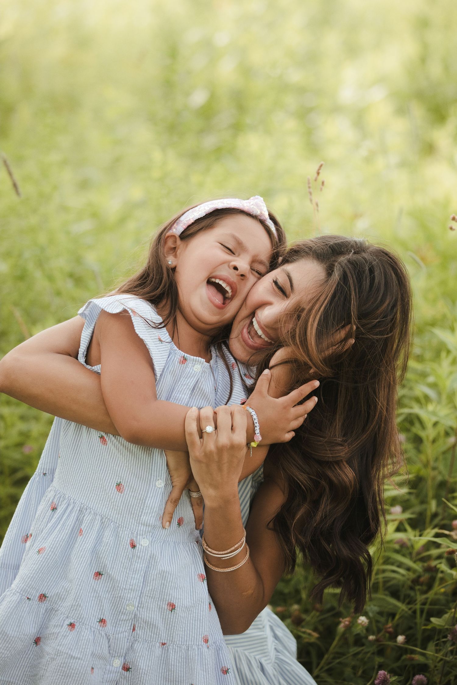 A joyful mother and daughter laughing and hugging in a lush green meadow, girl wearing a blue dress with a pink headband.