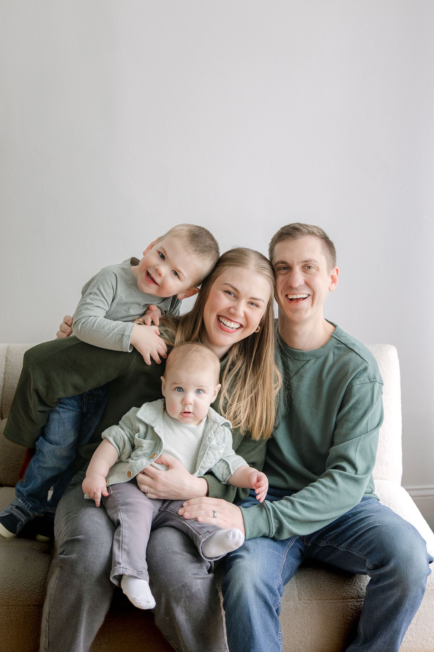 Happy family of four with two young children smiling together on a couch in a bright living room.
