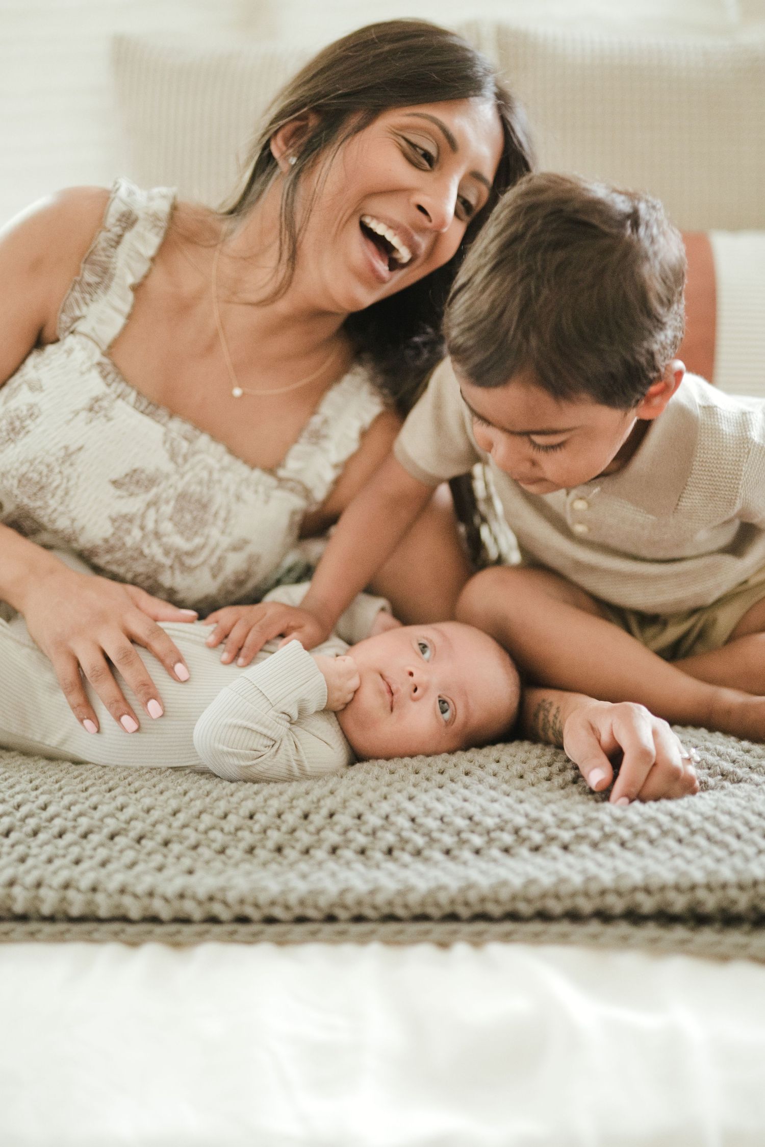 A laughing mother and young boy lovingly interact with a smiling newborn baby on a cozy knit blanket.