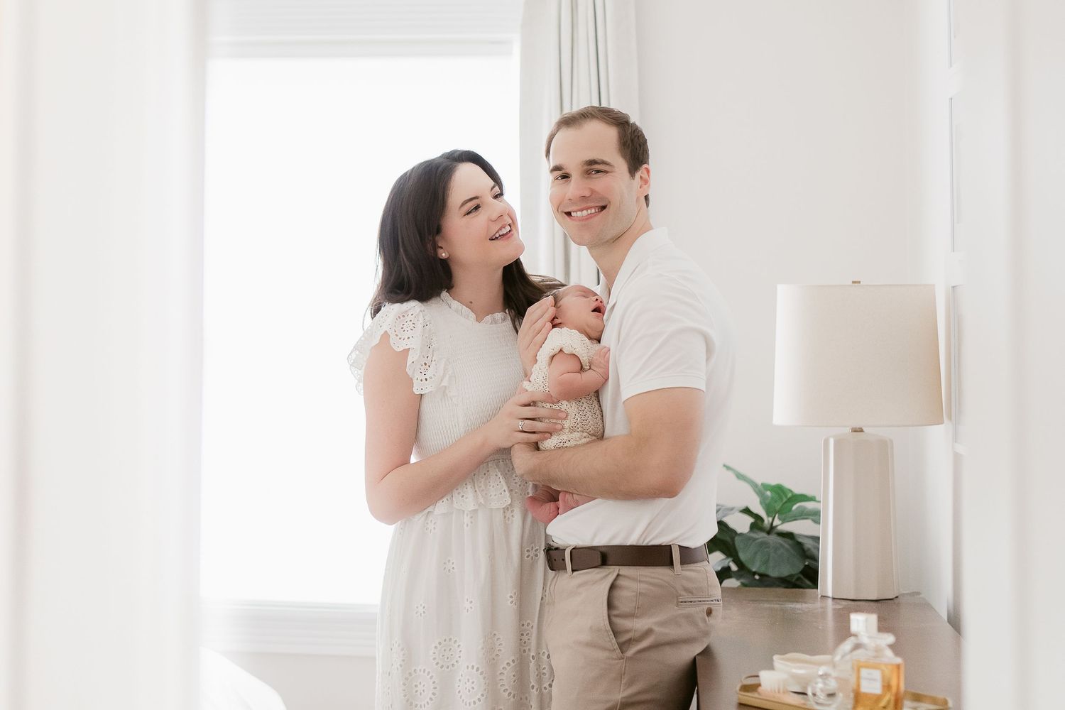 A couple in white stands together in a bright, airy room with natural light streaming through windows.