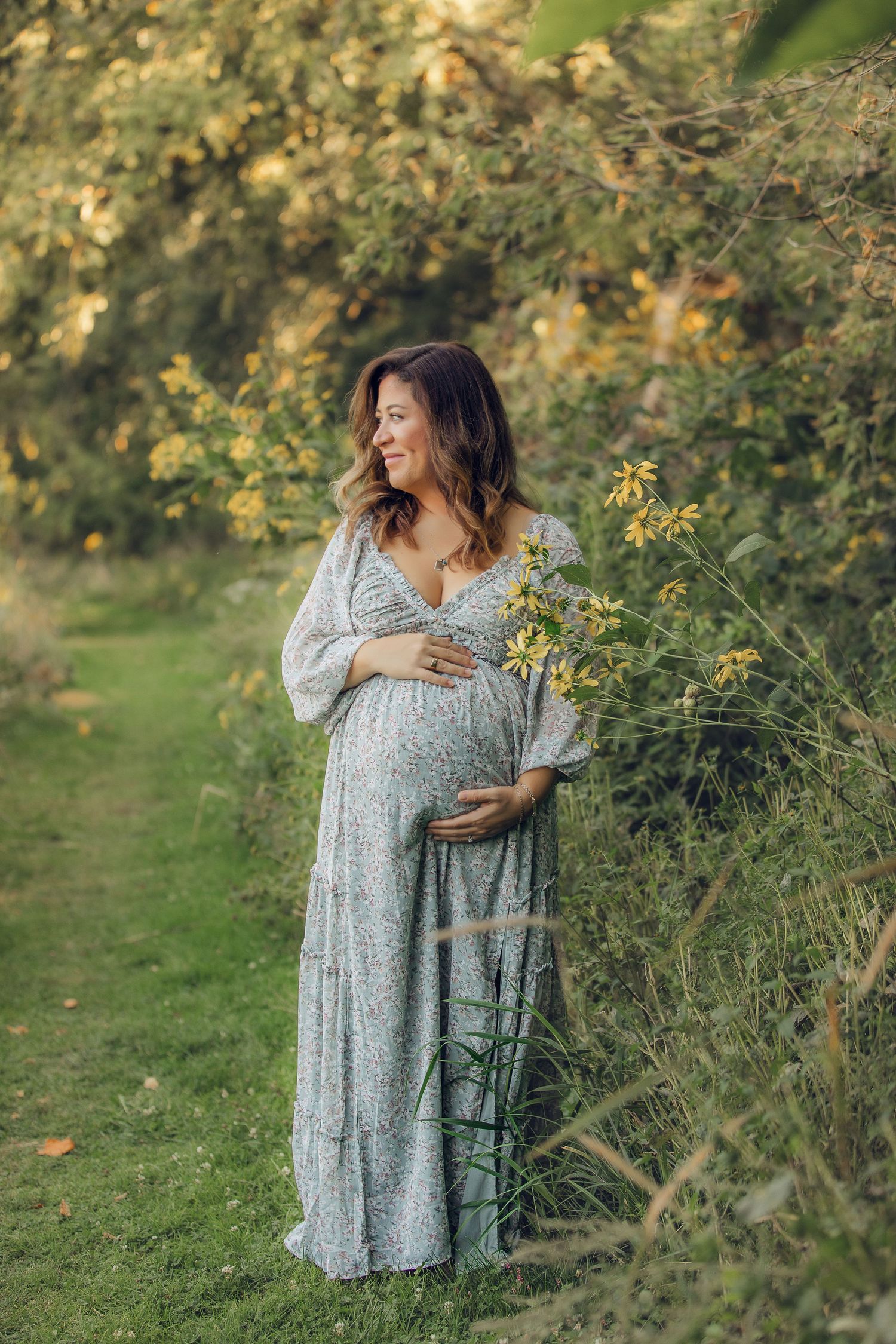 A woman in a floral dress poses for maternity photos in a garden setting with golden light.