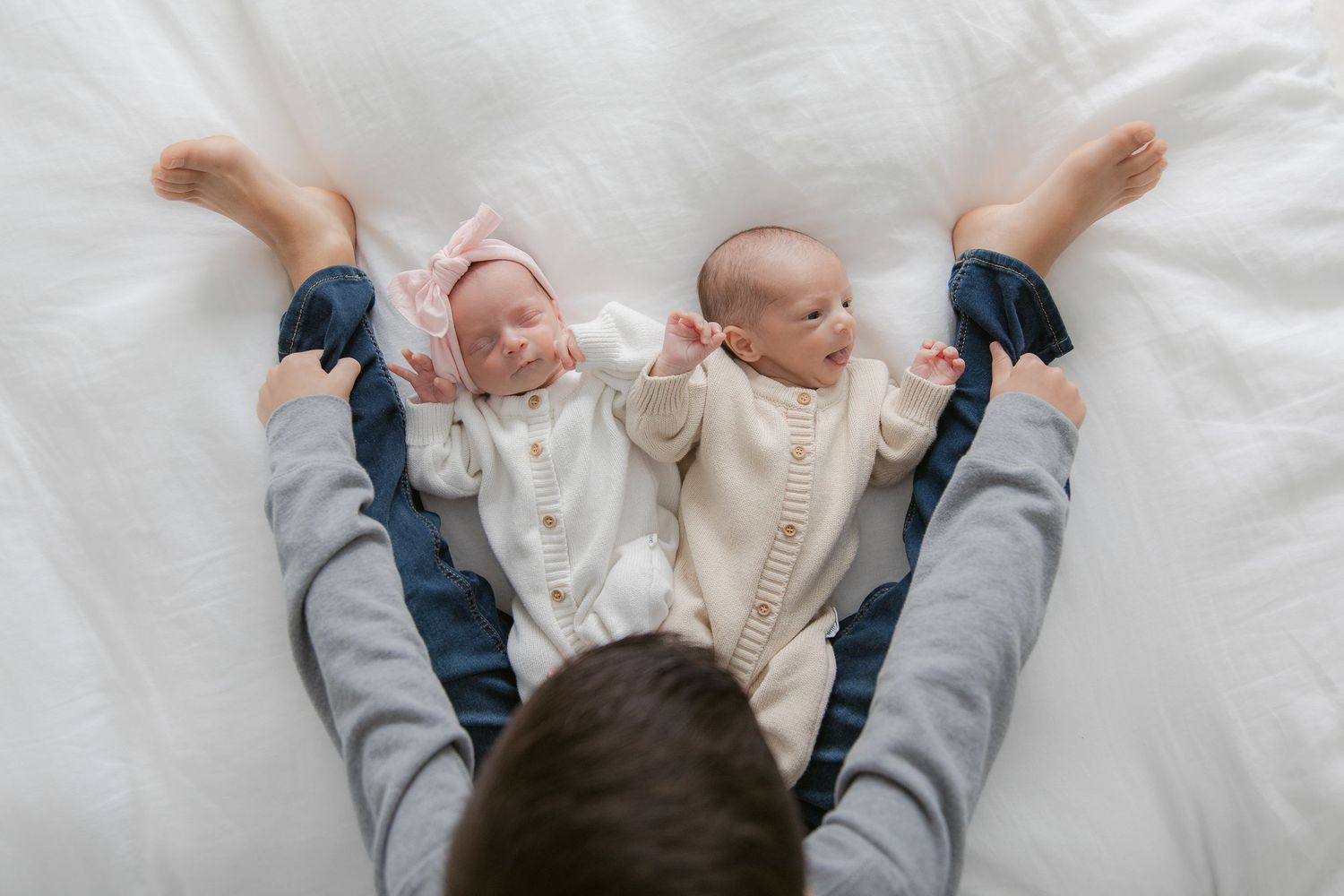 Newborn baby wearing white clothing lies on soft white bedding while stretching their legs upward.