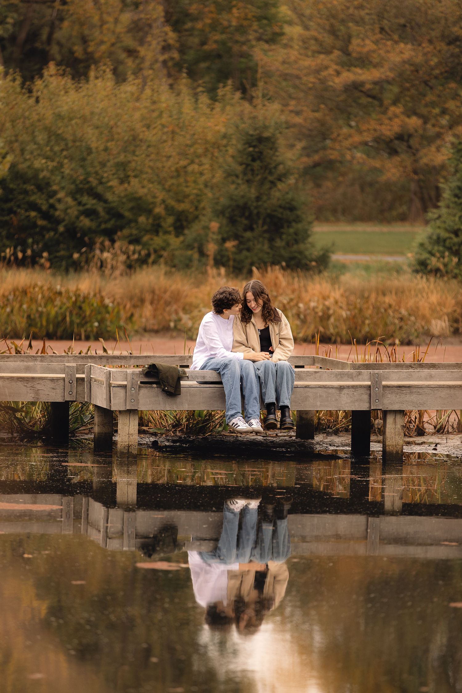 Couple sitting close together on a wooden dock by a calm reflective pond surrounded by autumn foliage.