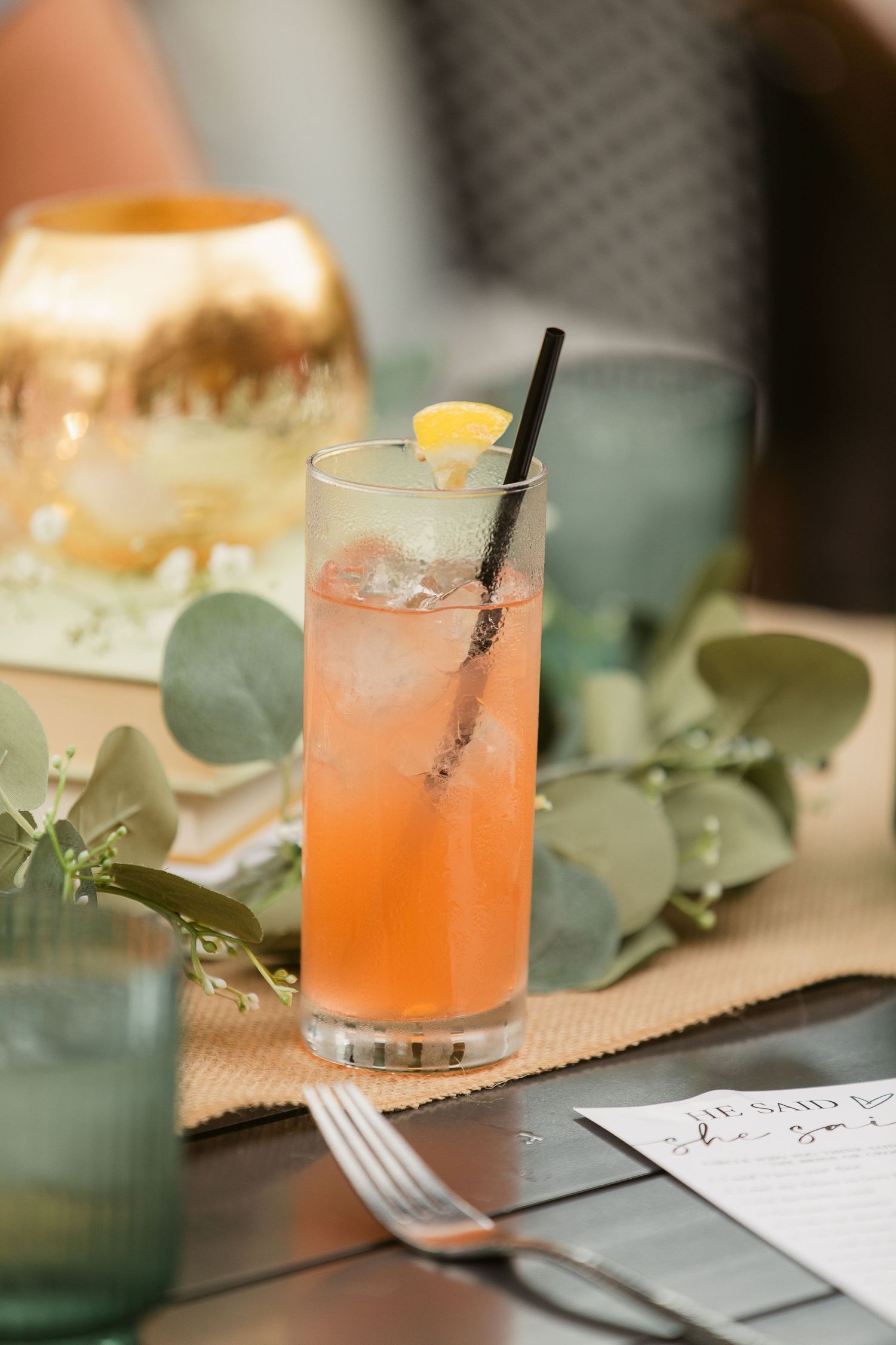 A pink cocktail with ice, black straw, and lemon garnish on a table with eucalyptus leaves and golden decor.