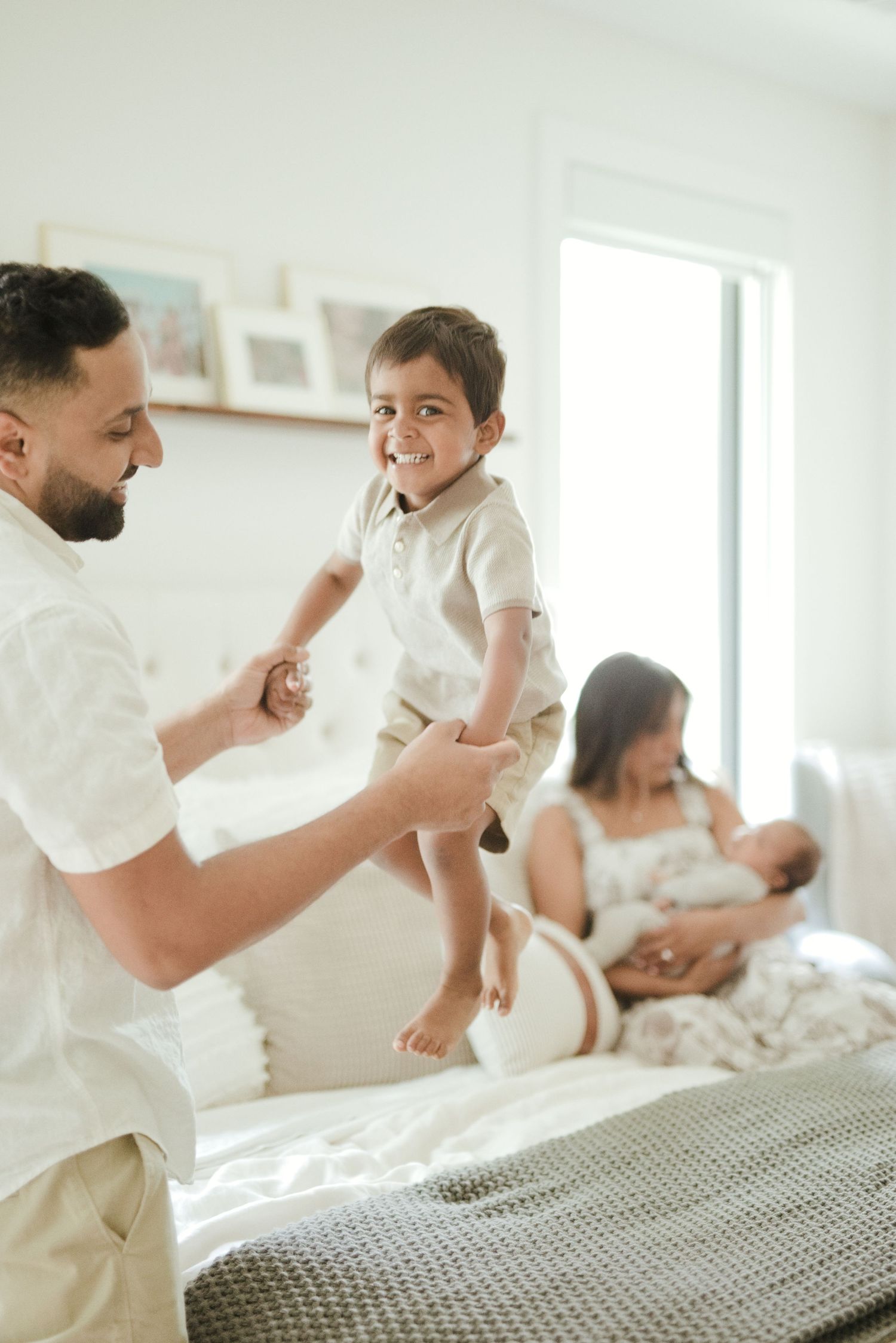 A father lifts his smiling toddler son on a white bed while mother sits behind nursing a newborn baby.