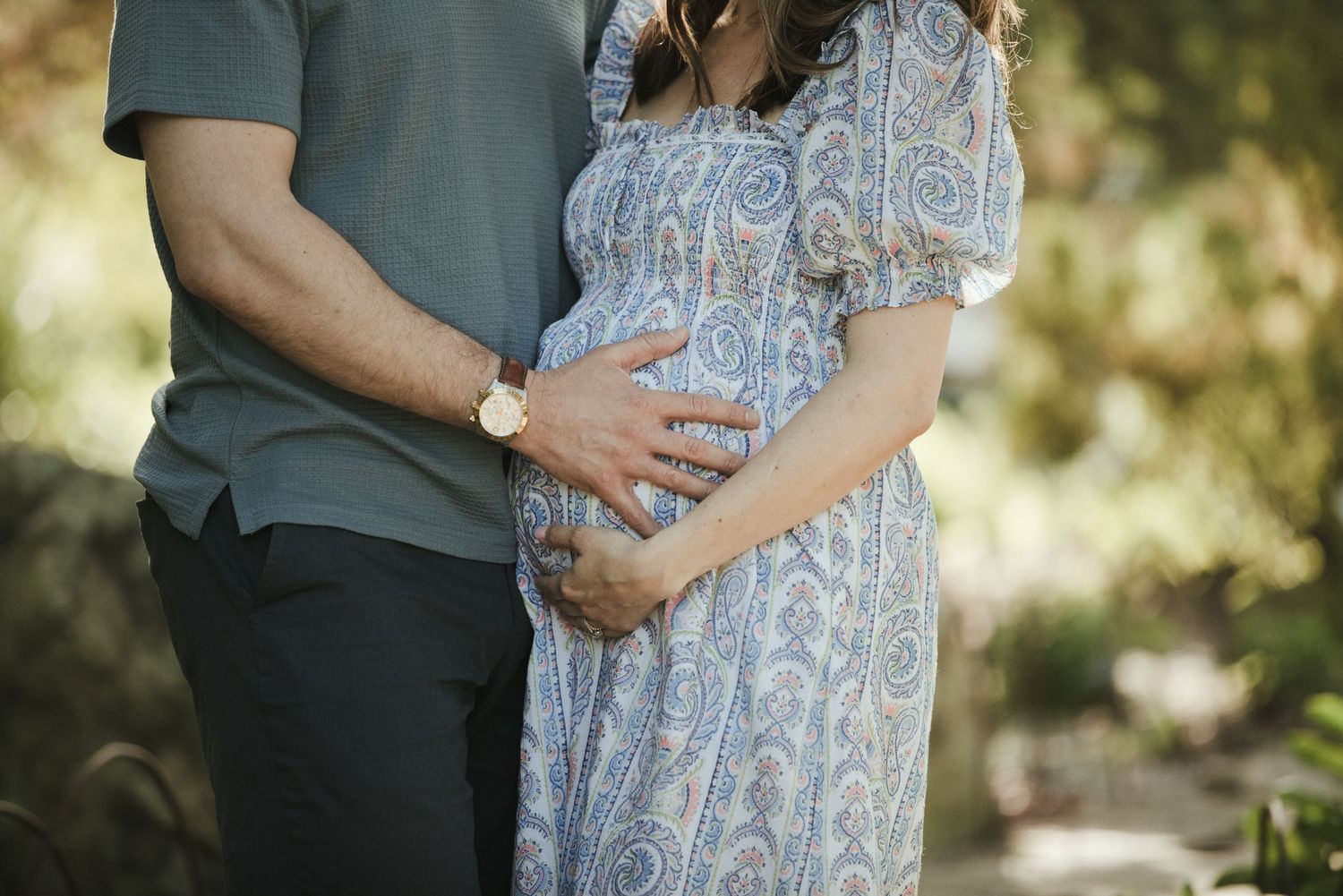 Couple tenderly cradling pregnant belly outdoors, woman in blue paisley maternity dress, man in teal shirt.