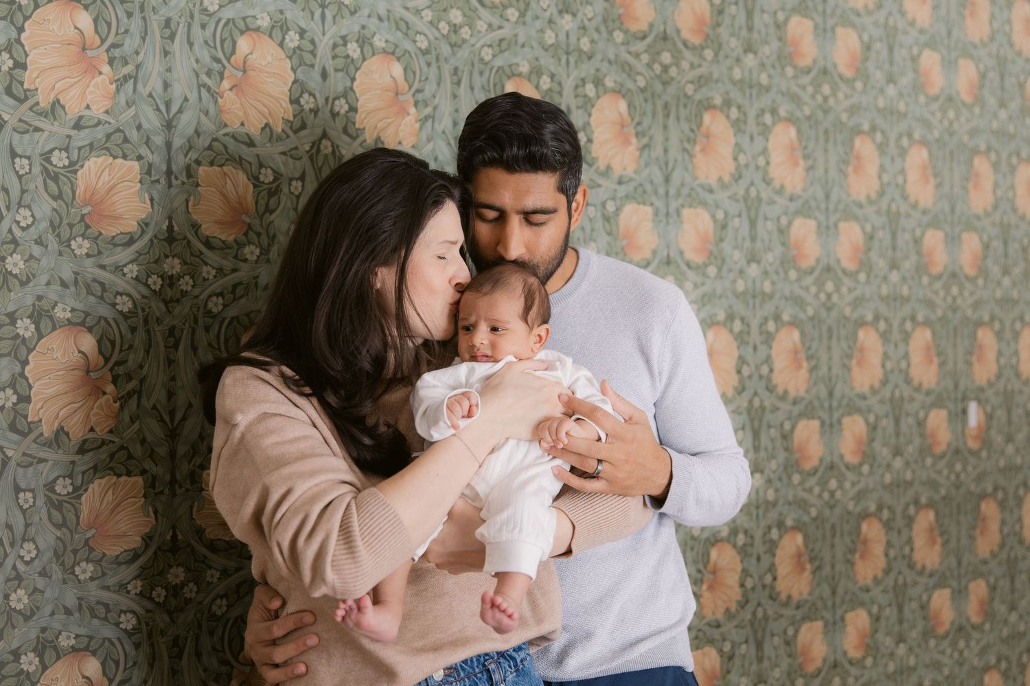 Parents cradle their newborn baby together against a decorative patterned wall background.