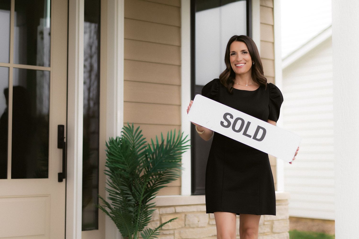 Person in black dress holding Sold sign outside modern home entrance.