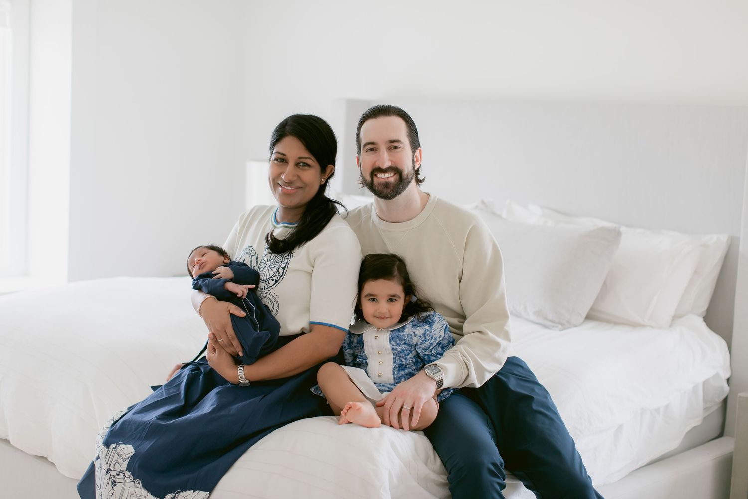 Happy family of four posing on a white bed, parents with newborn baby and young daughter smiling.