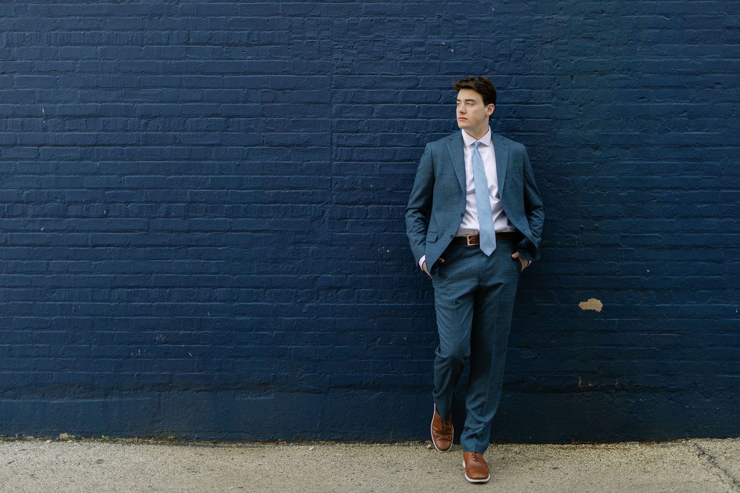 A young man in a blue suit and tie leans against a dark navy brick wall with hands in pockets.
