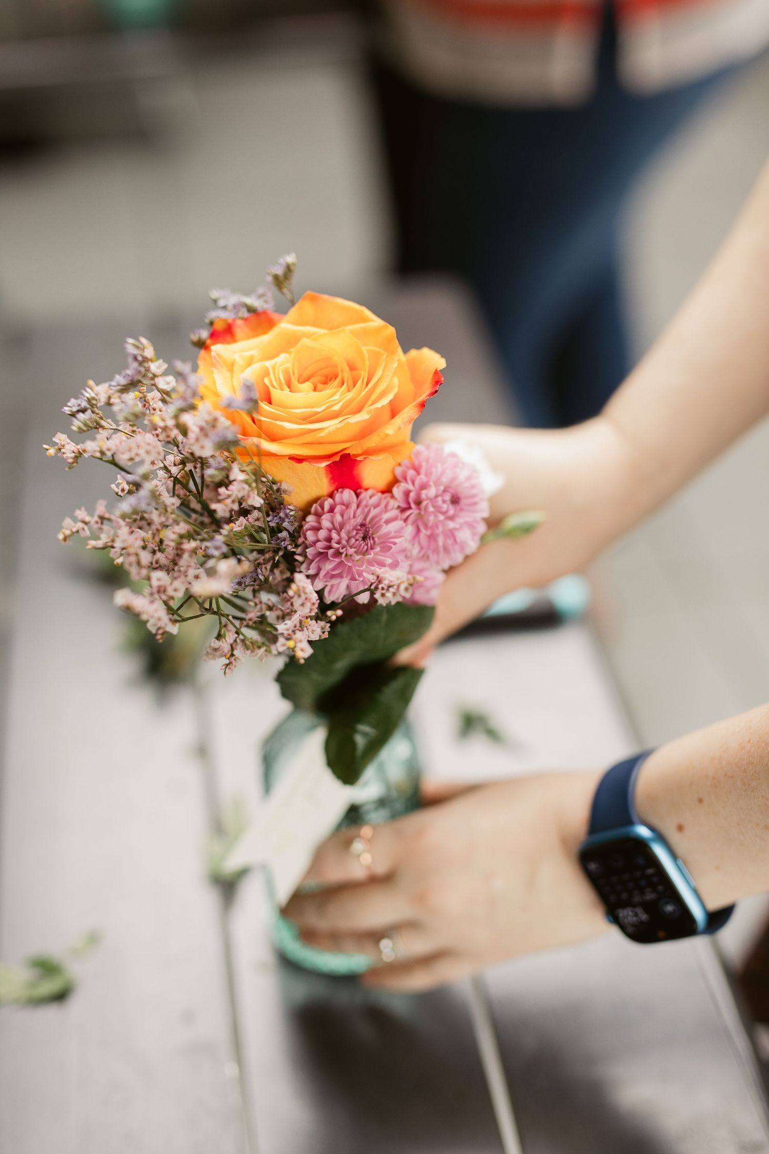Florist arranging a vibrant bouquet with orange rose and purple flowers in a glass mason jar vase.