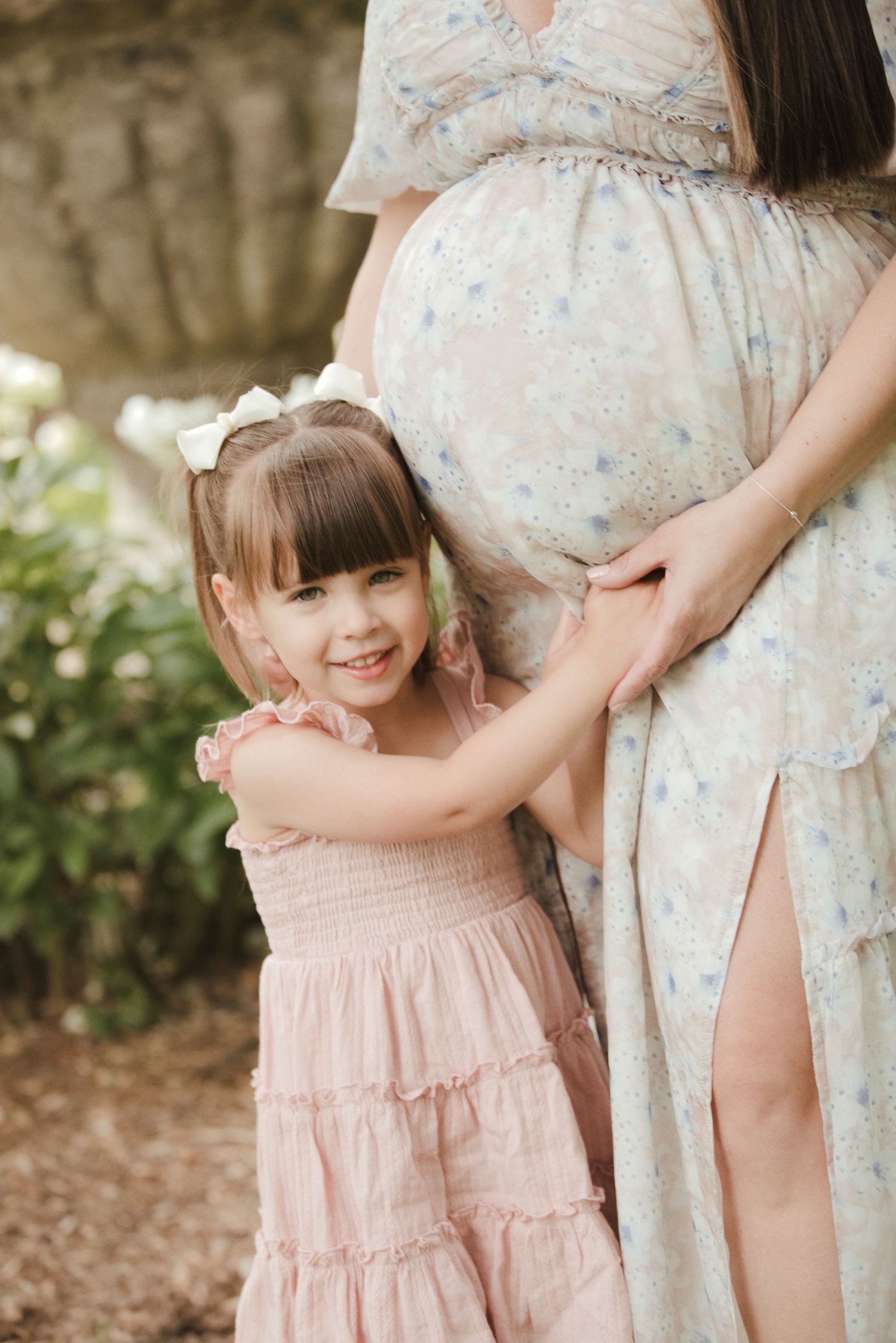 A young girl in a pink dress hugs her pregnant mother's belly outdoors during a maternity photo session.