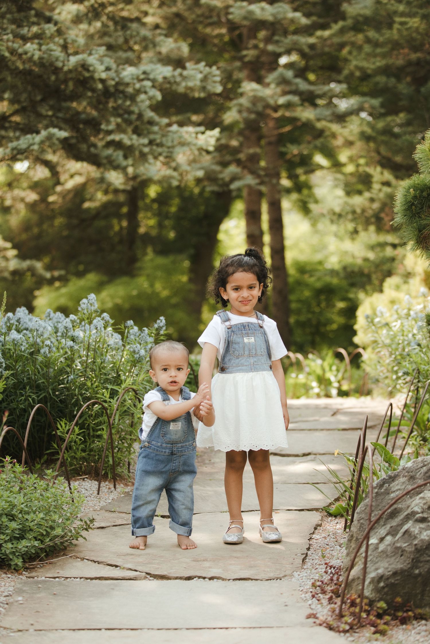 Two young siblings holding hands on a garden path, older girl in denim overalls and white dress, toddler in jeans outdoors.