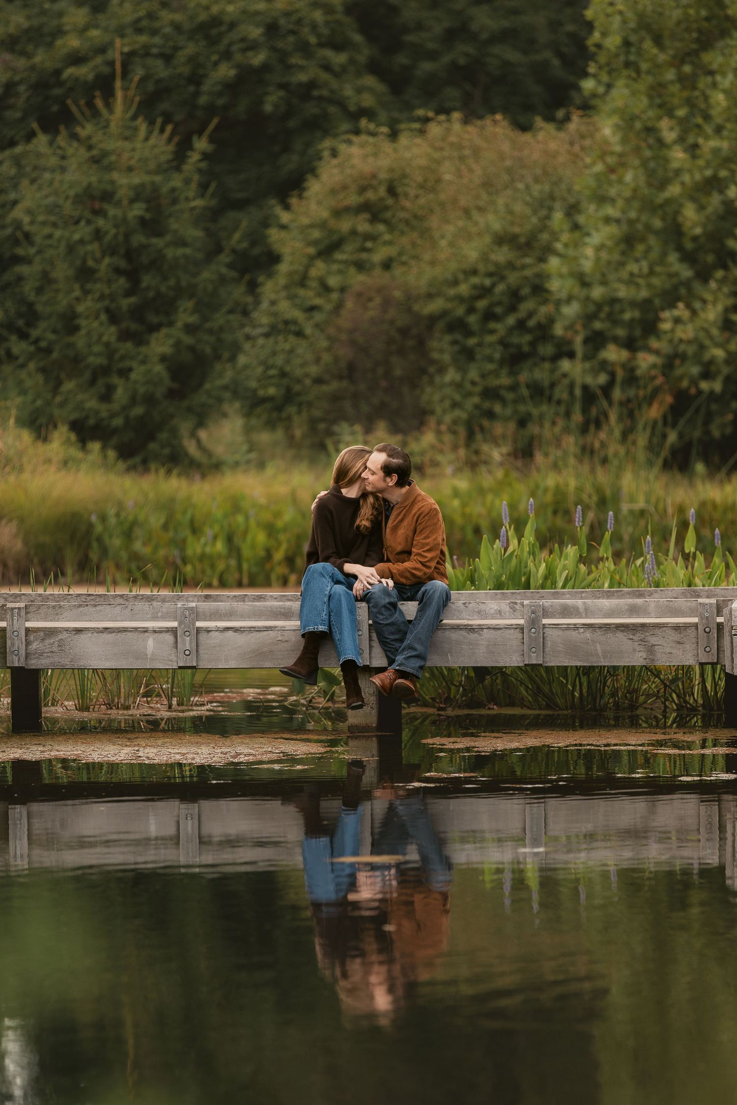 Couple sharing a romantic moment on a wooden dock by a reflective pond, surrounded by lush green trees.