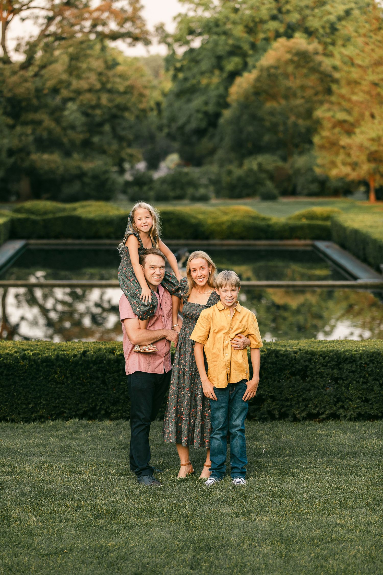 Family of four posing in a lush garden with a reflecting pool, parents holding children, wearing warm autumn colors.