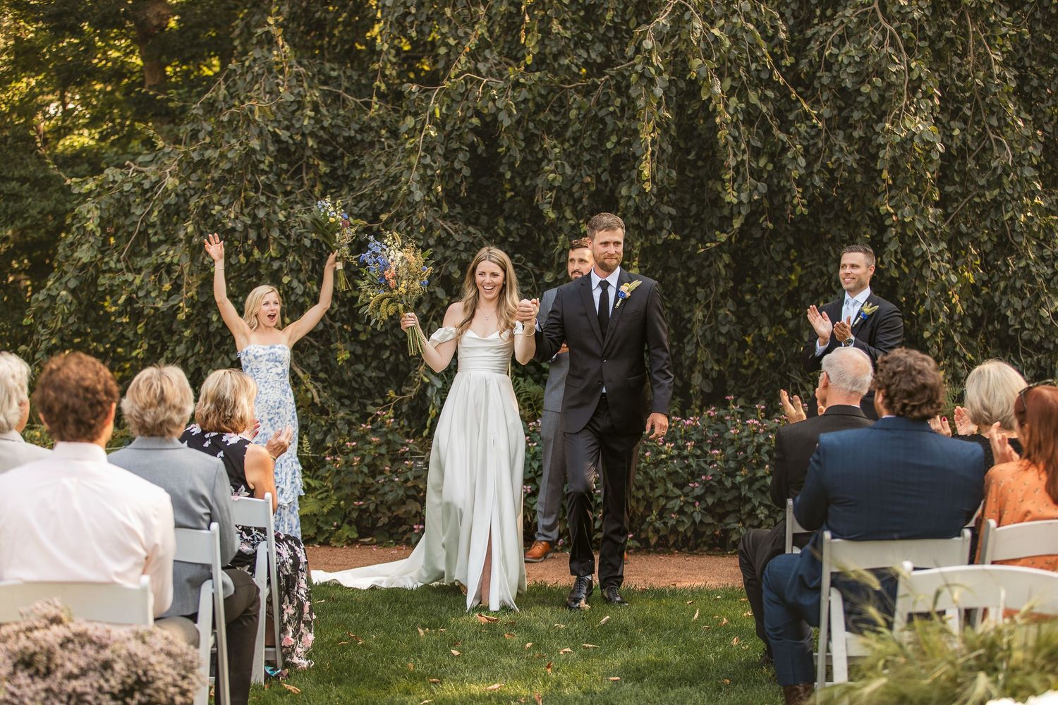Joyful celebration at an outdoor garden wedding ceremony with guests seated in white chairs and greenery backdrop.