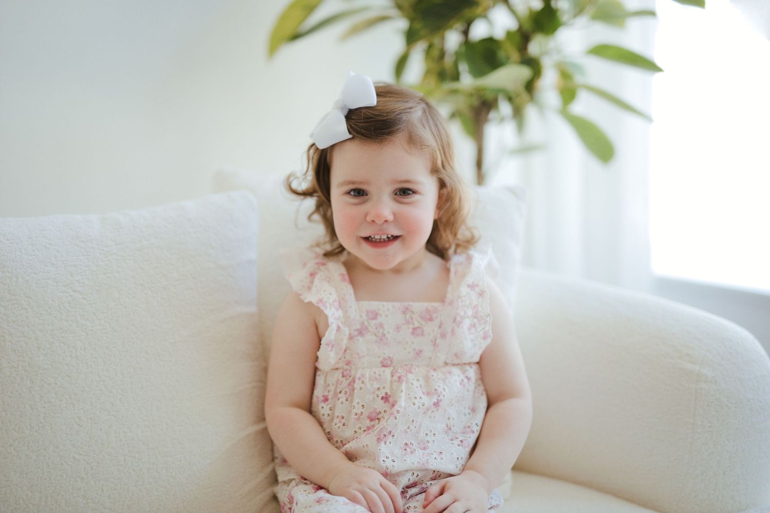 Smiling toddler girl in floral dress with white bow sits on white sofa.