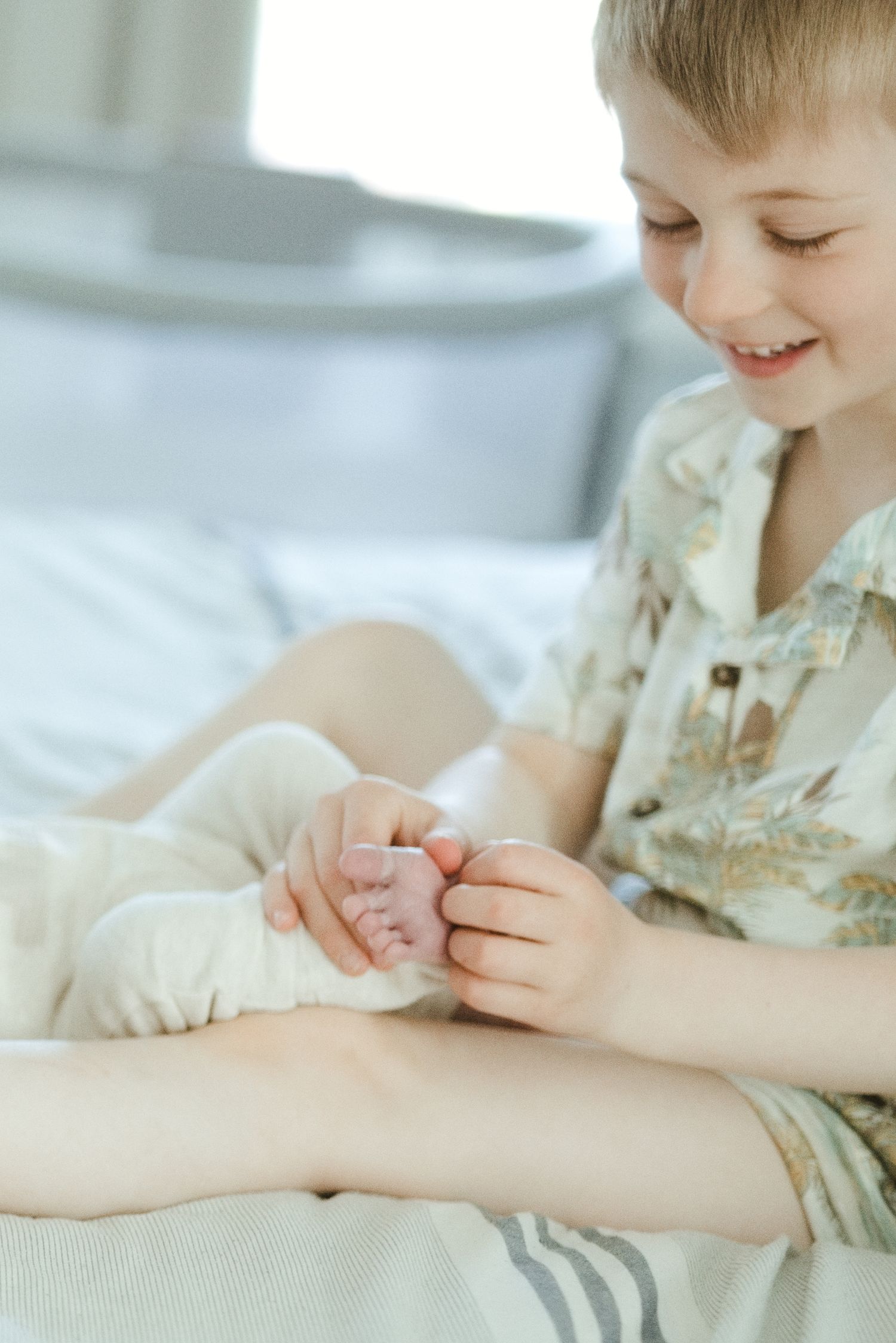 Smiling young boy sitting on a bed holding a baby's tiny feet, wearing a floral shirt.