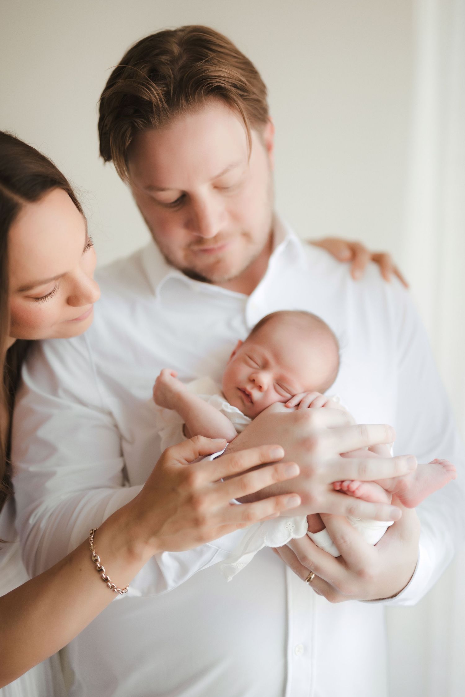 New parents in white shirts tenderly hold their sleeping newborn baby together in a bright, minimalist setting.