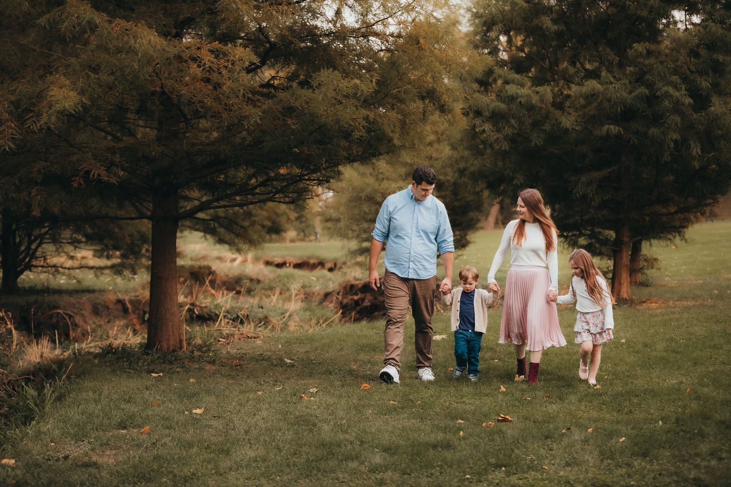 A family walks hand in hand through a grassy park on an autumn afternoon with trees creating a warm backdrop.
