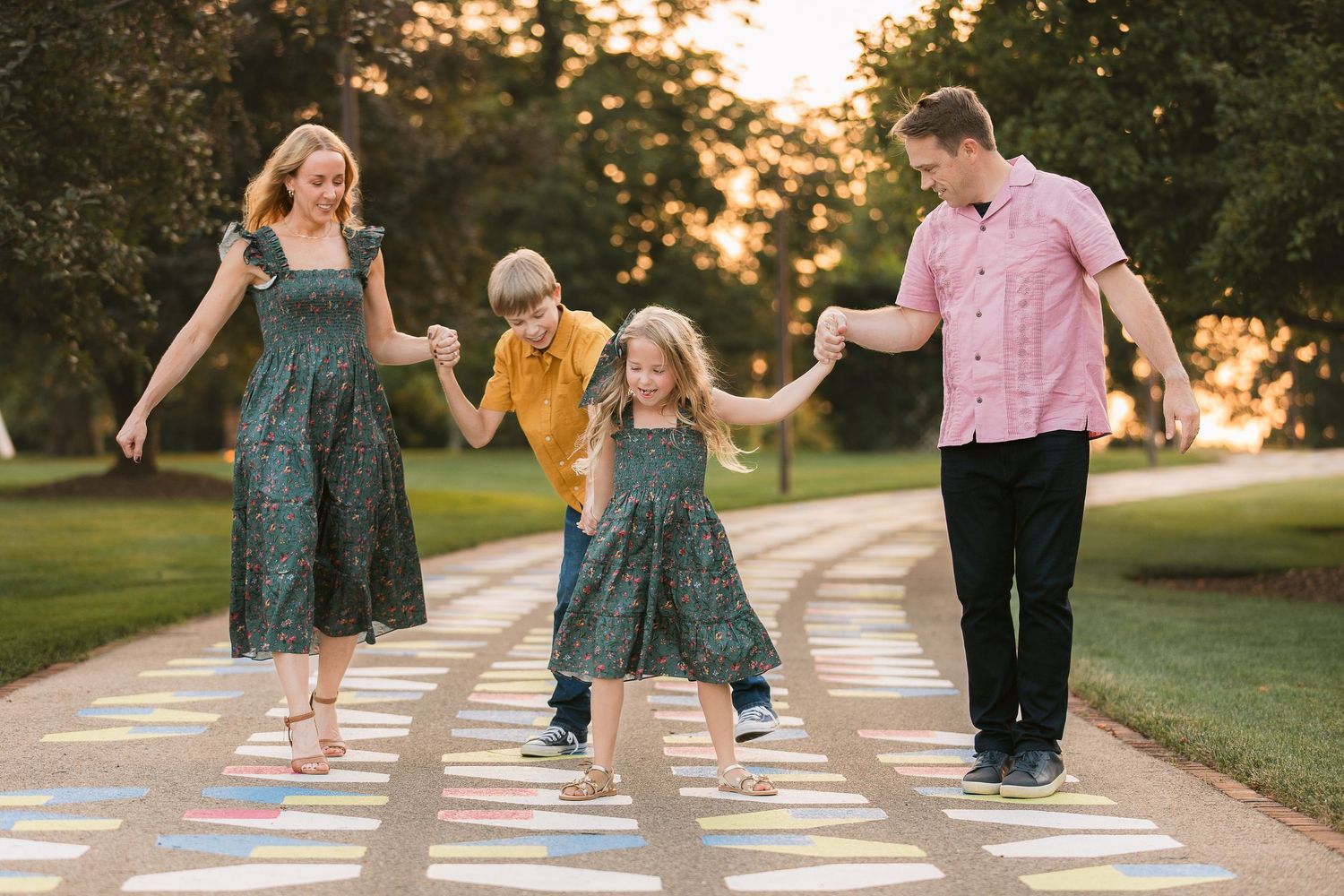 Family laughs and plays together during a creative sidewalk chalk art game at sunset on a neighborhood pathway.