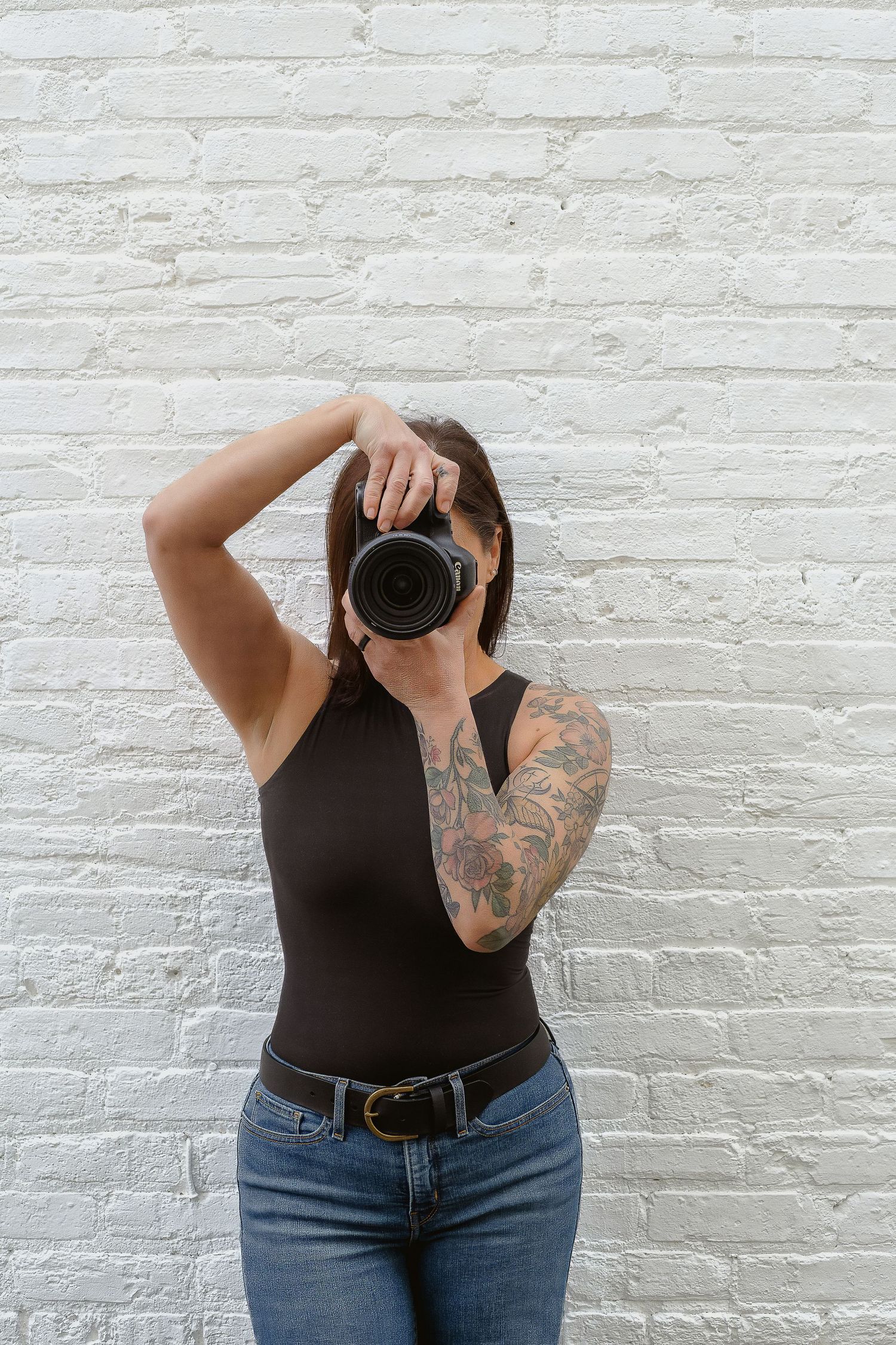A tattooed woman in a black bodysuit and jeans holds a DSLR camera up to her face against a white brick wall.