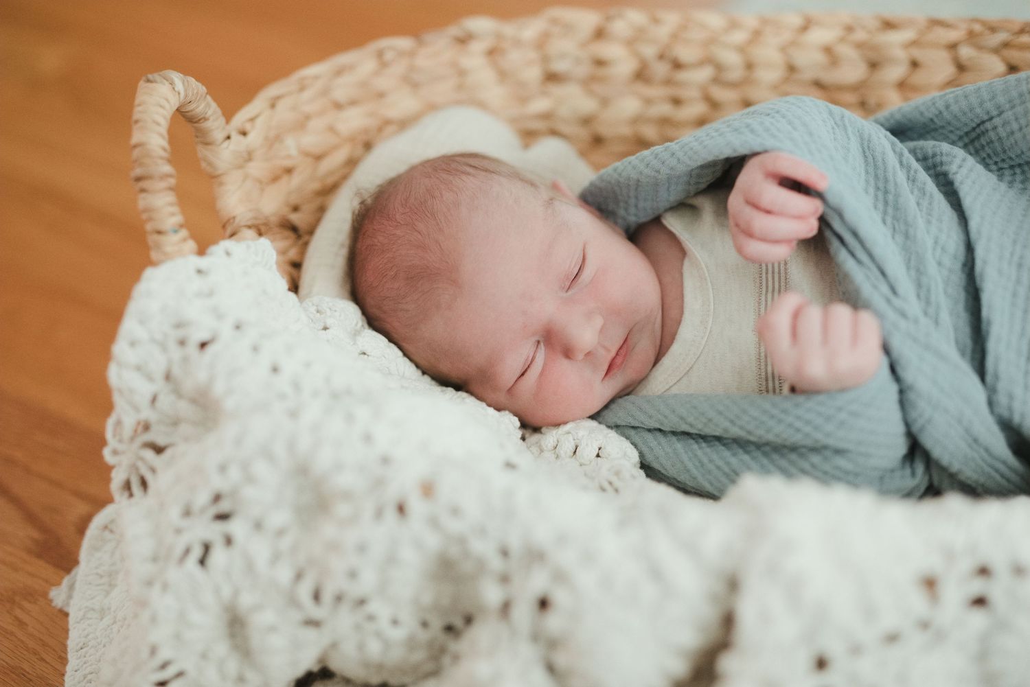Peaceful newborn baby sleeping in a wicker basket wrapped in a soft teal blanket on a white knit blanket.