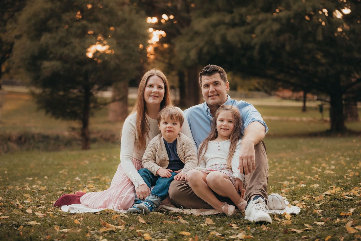 Family sitting together on autumn ground covered in fallen leaves during golden hour sunset in park setting.