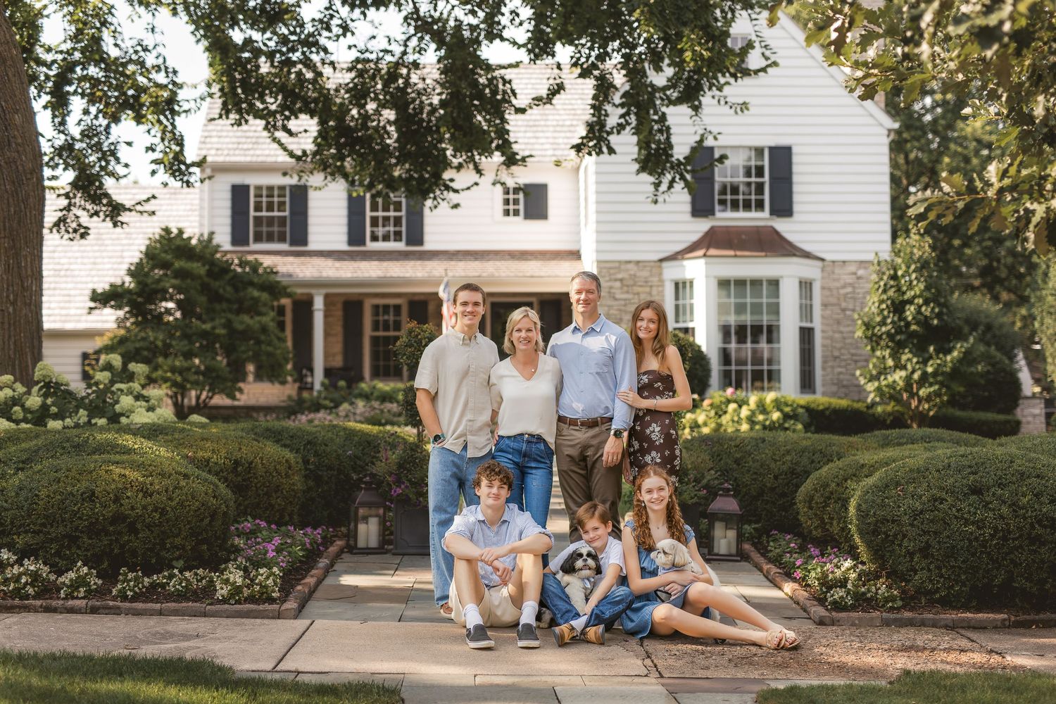 Multi-panel collage of lifestyle family portraits outdoors near traditional white colonial style homes and gardens.