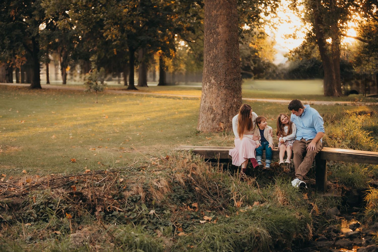 People relaxing on a wooden bench in a sunlit park during golden hour with trees and grass in the background.