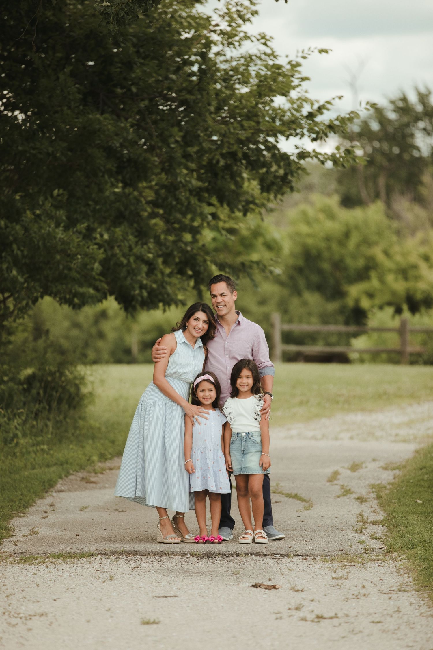 A happy family of four poses on a gravel path surrounded by lush green trees during an outdoor photo session.