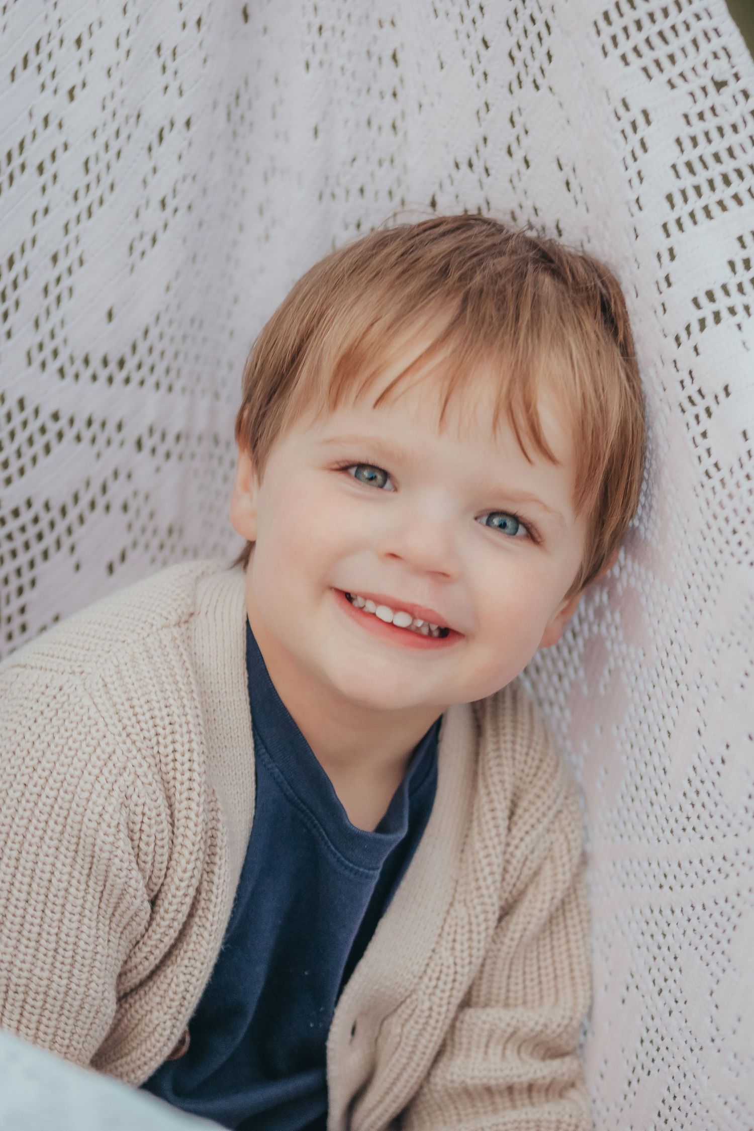 Child wearing a beige cardigan and navy shirt smiles joyfully while posing against a white lace backdrop.