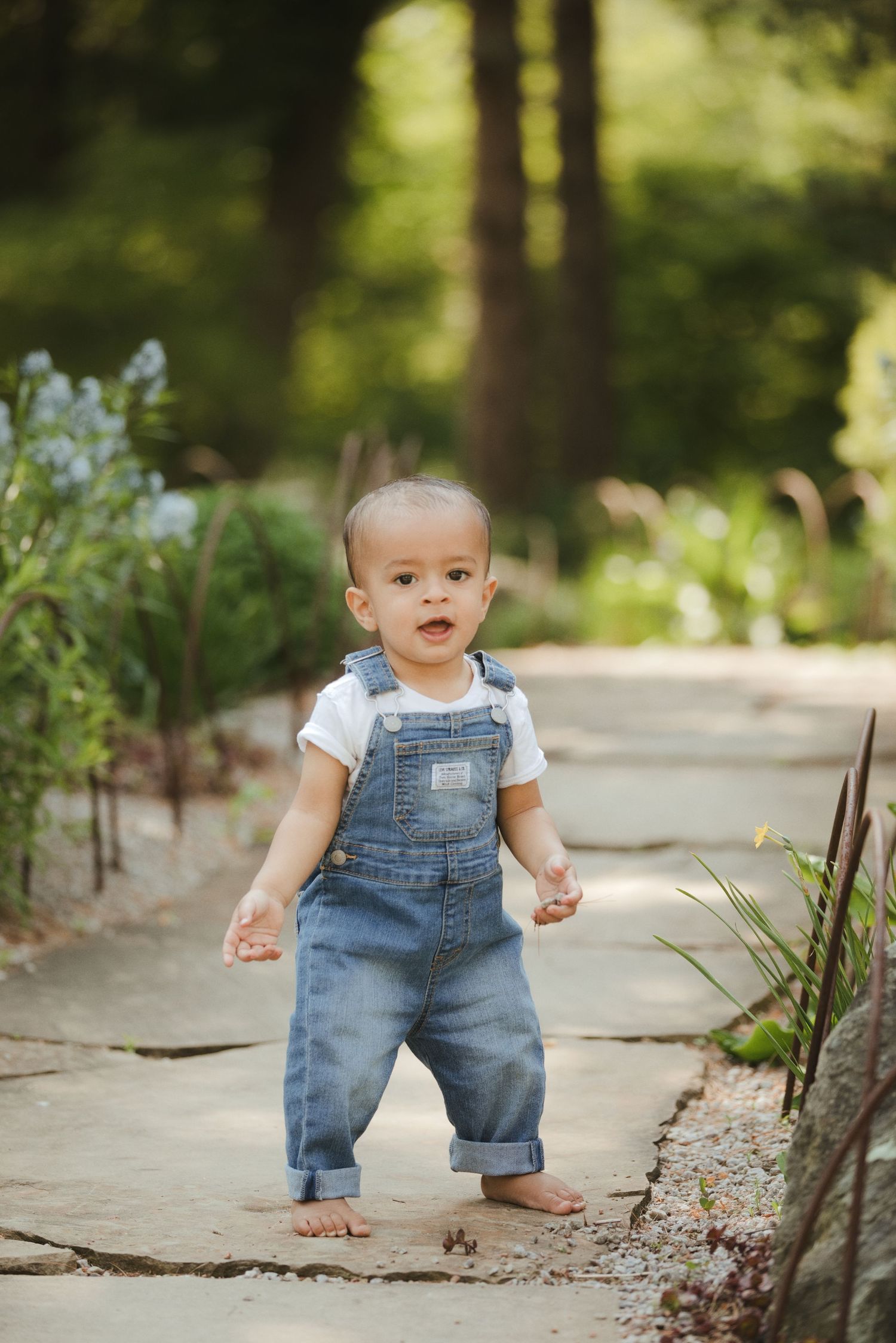 Adorable toddler in denim overalls taking first steps on a garden path surrounded by greenery.