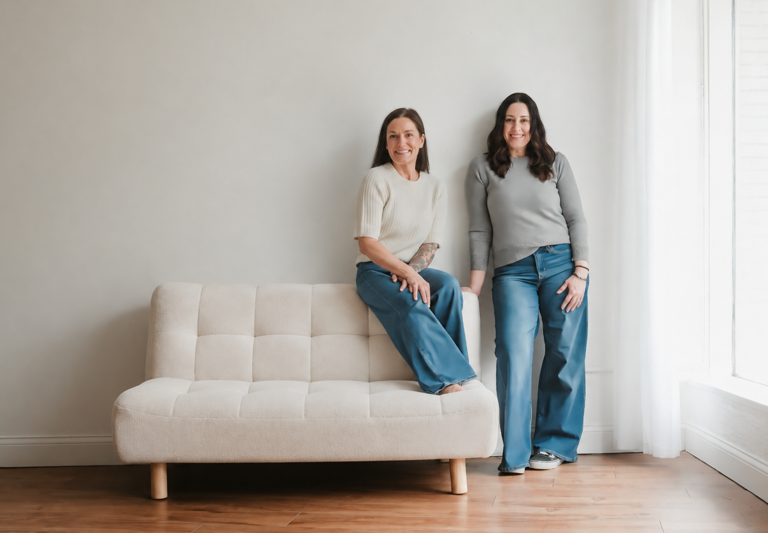 Two women smiling in jeans pose beside a white tufted sofa on hardwood floors against a light gray wall.
