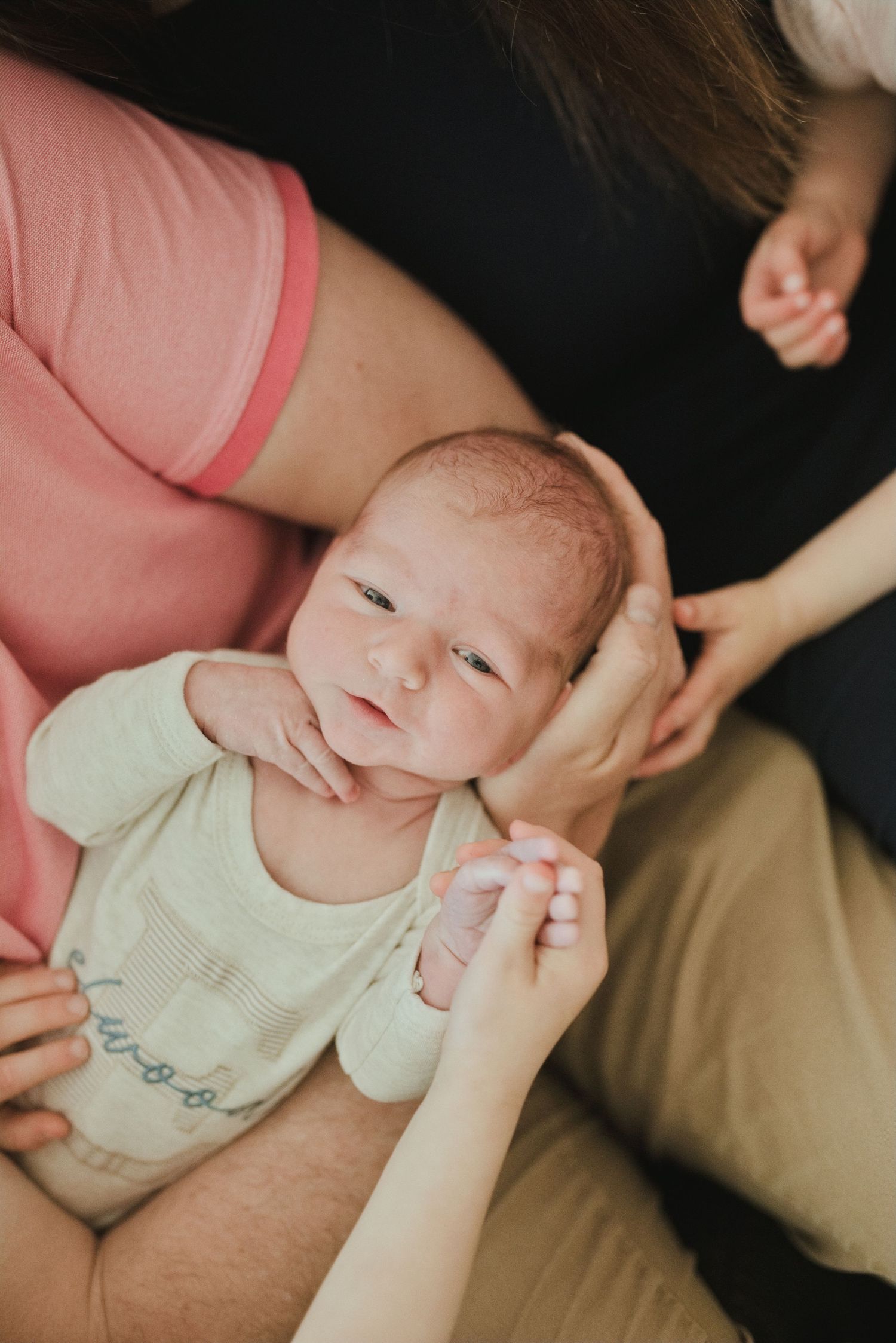 Newborn baby in white onesie cradled in parent's arms, looking up with alert eyes during a tender family moment.