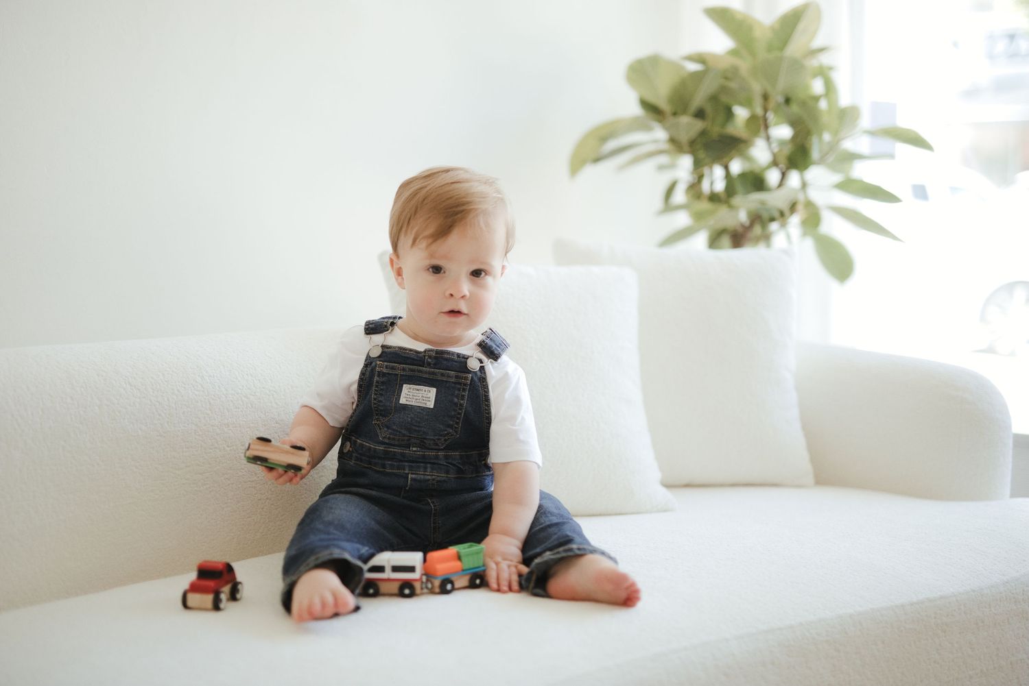 Baby in denim overalls sitting on white sofa playing with toy cars in bright living room.