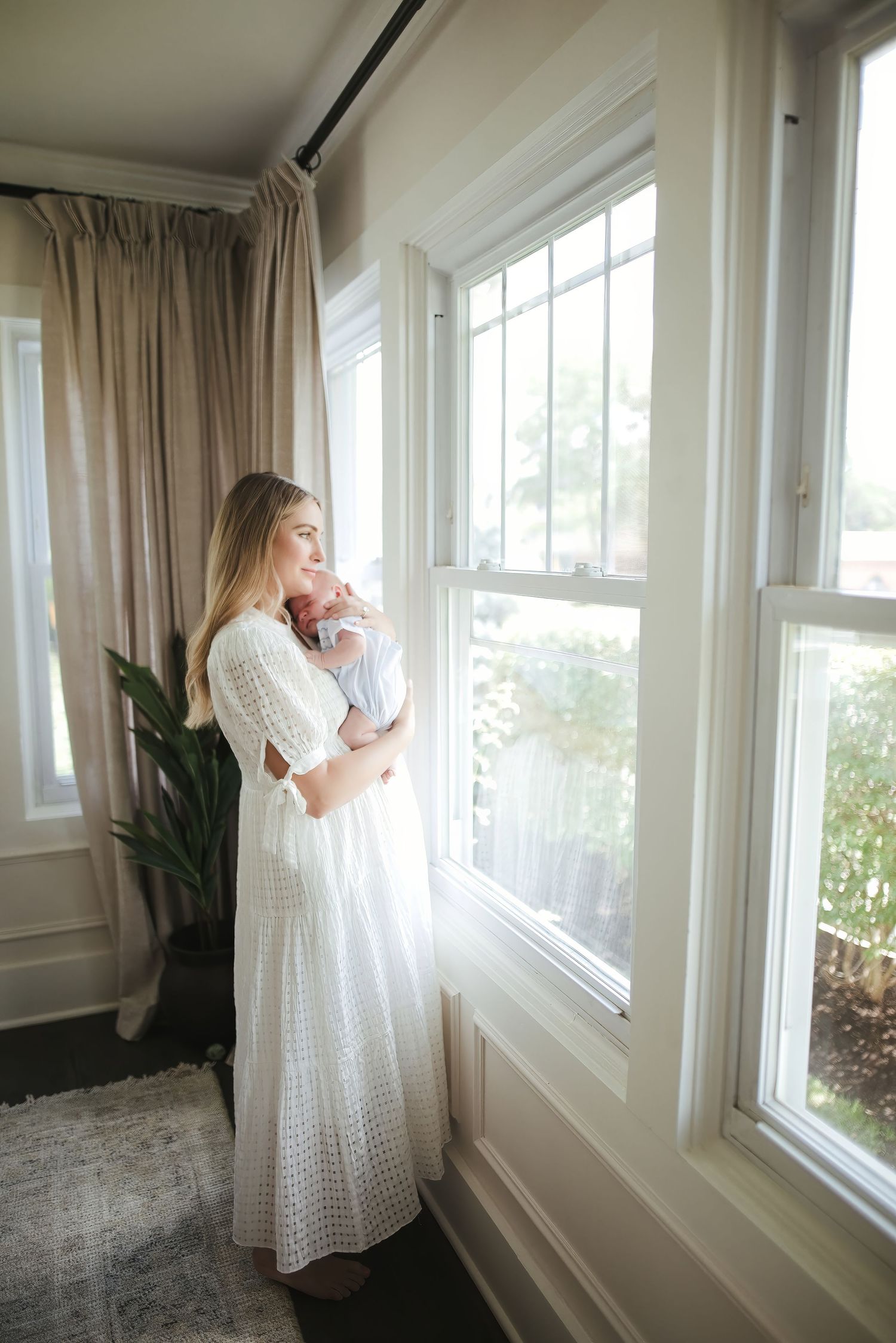 Woman in white dress holding newborn baby by sunlit window in elegant home interior.