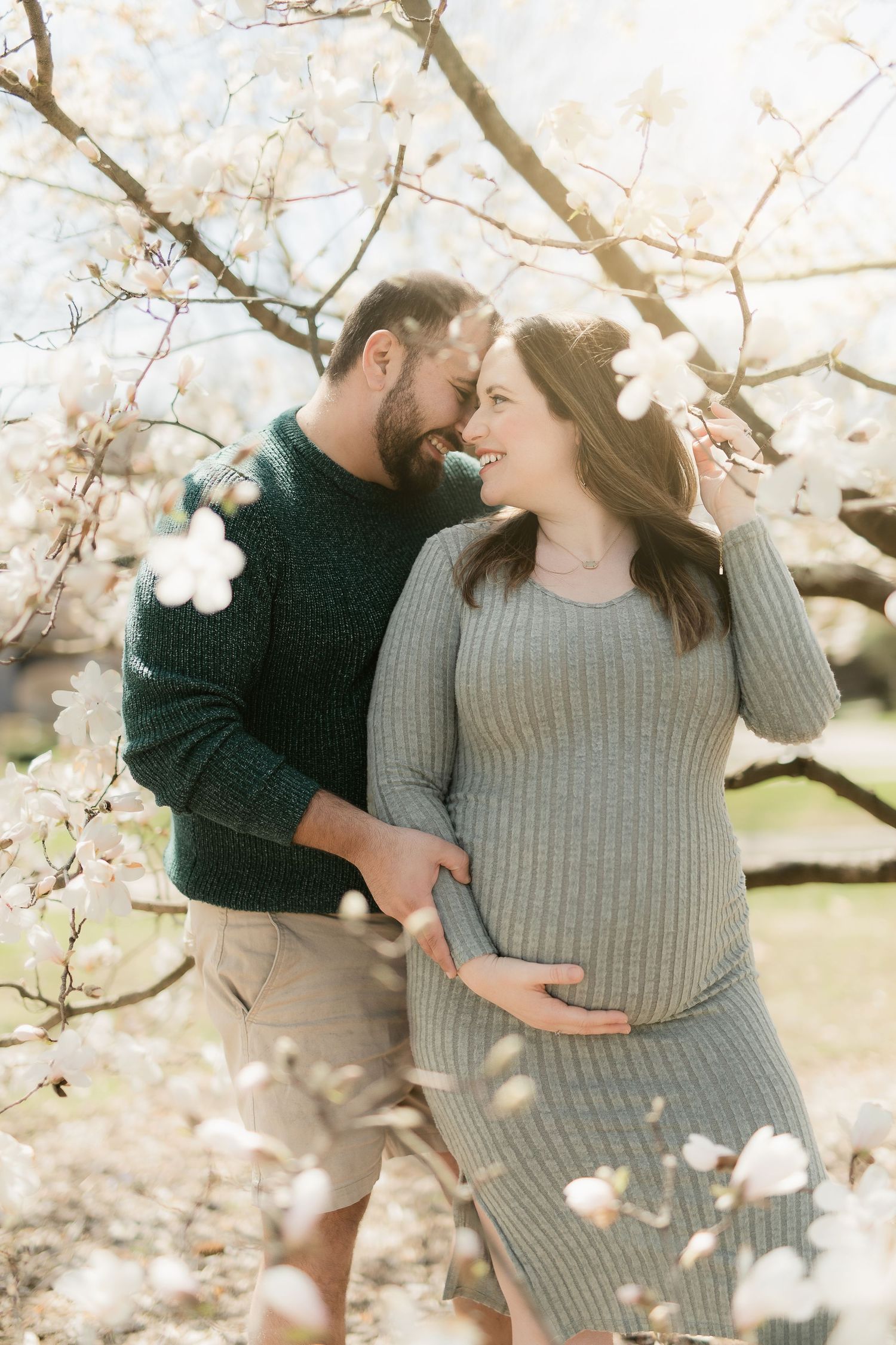 A couple embraces under flowering cherry blossoms as they celebrate their pregnancy in a romantic outdoor photo.