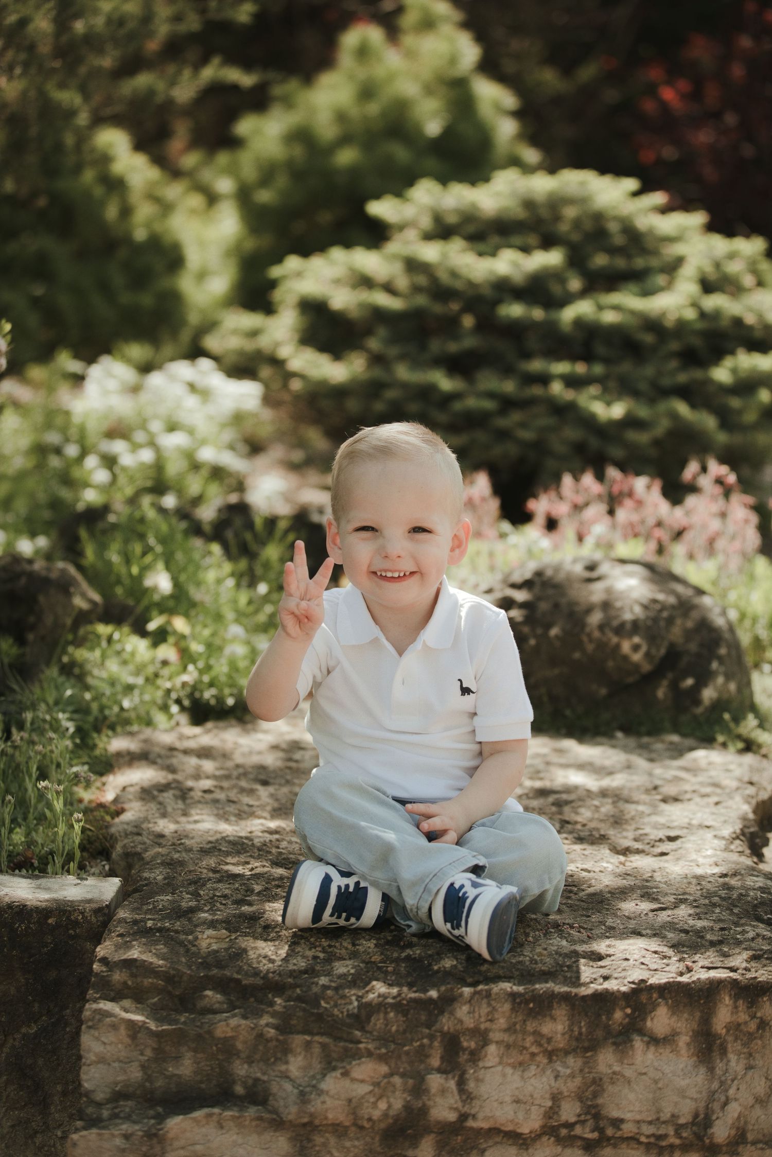 Smiling toddler boy in white polo and jeans sits on stone wall flashing peace sign in lush garden setting.
