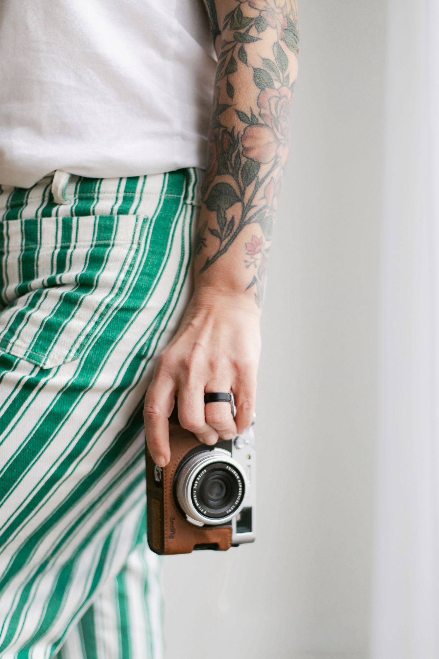 Person wearing green striped pants holds a vintage camera while showing floral arm tattoos and black ring.