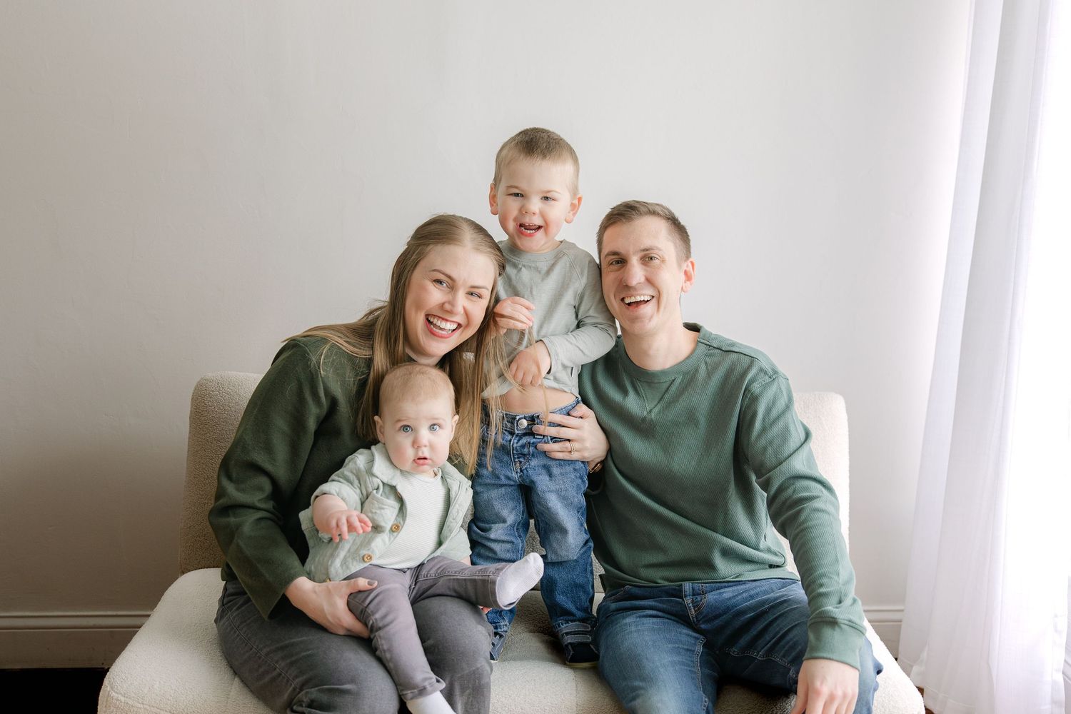 A family sits together on a couch in a white room wearing coordinated green and gray outfits.
