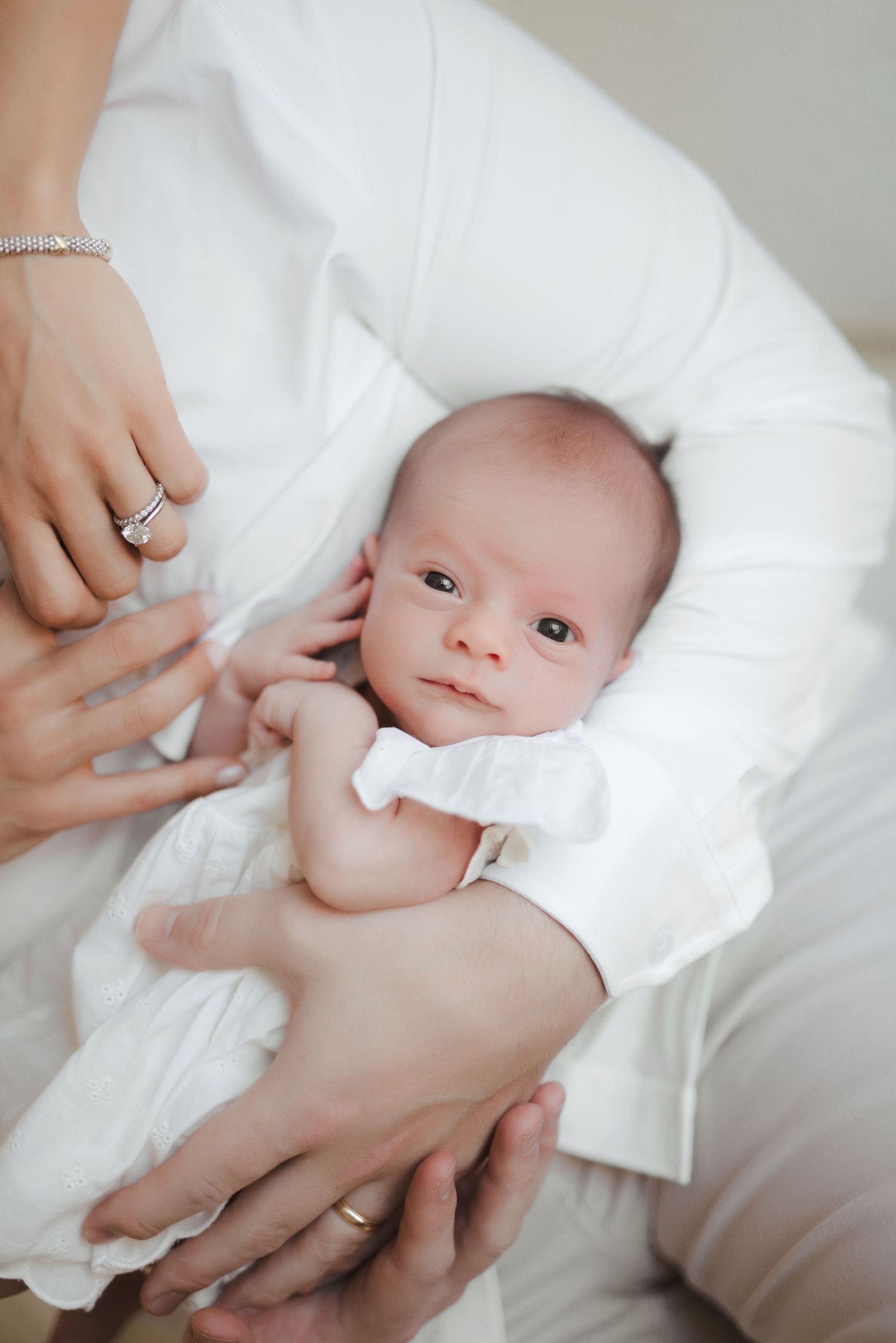 Newborn baby in white outfit cradled in mother's arms, with a calm expression and tiny hands visible.