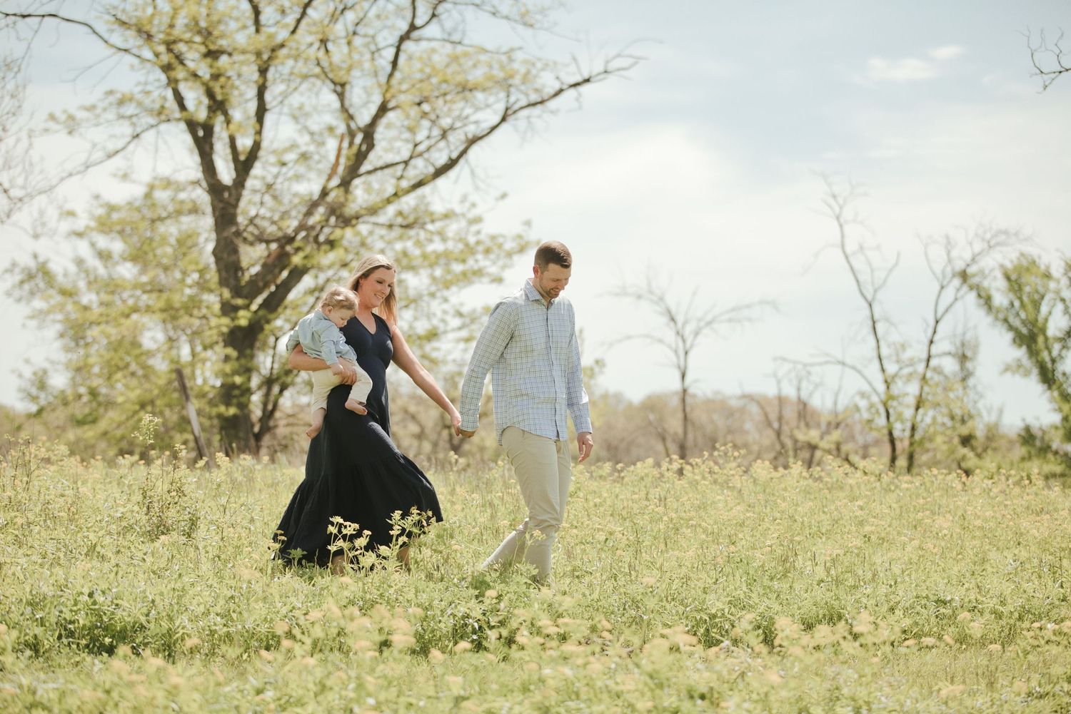 A couple holds hands and walks through a blooming meadow, the woman in a flowing black dress and the man in a light blue shirt.