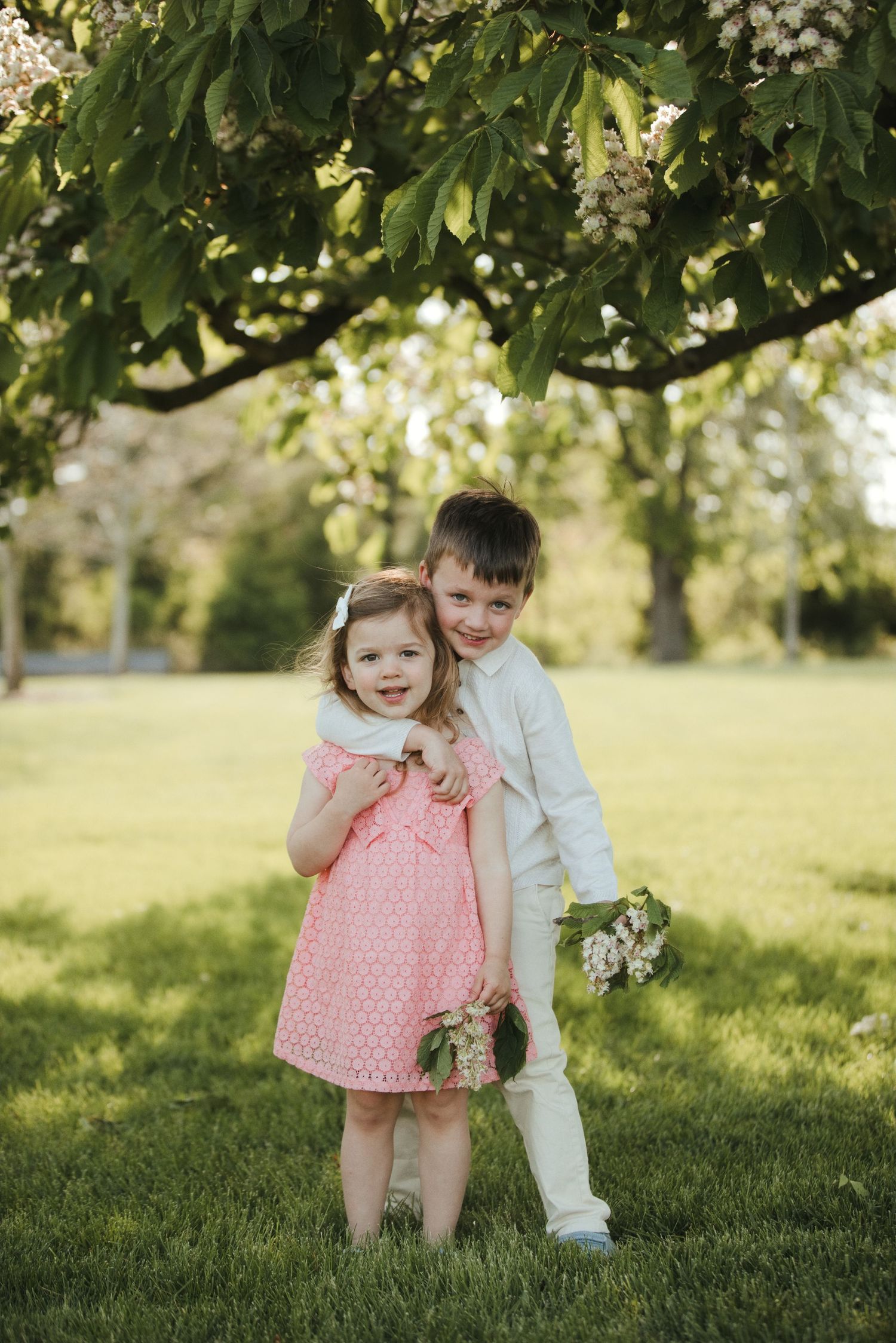Two young children pose under a blossoming tree in a green park, girl in pink dress, boy in white suit holding flowers.